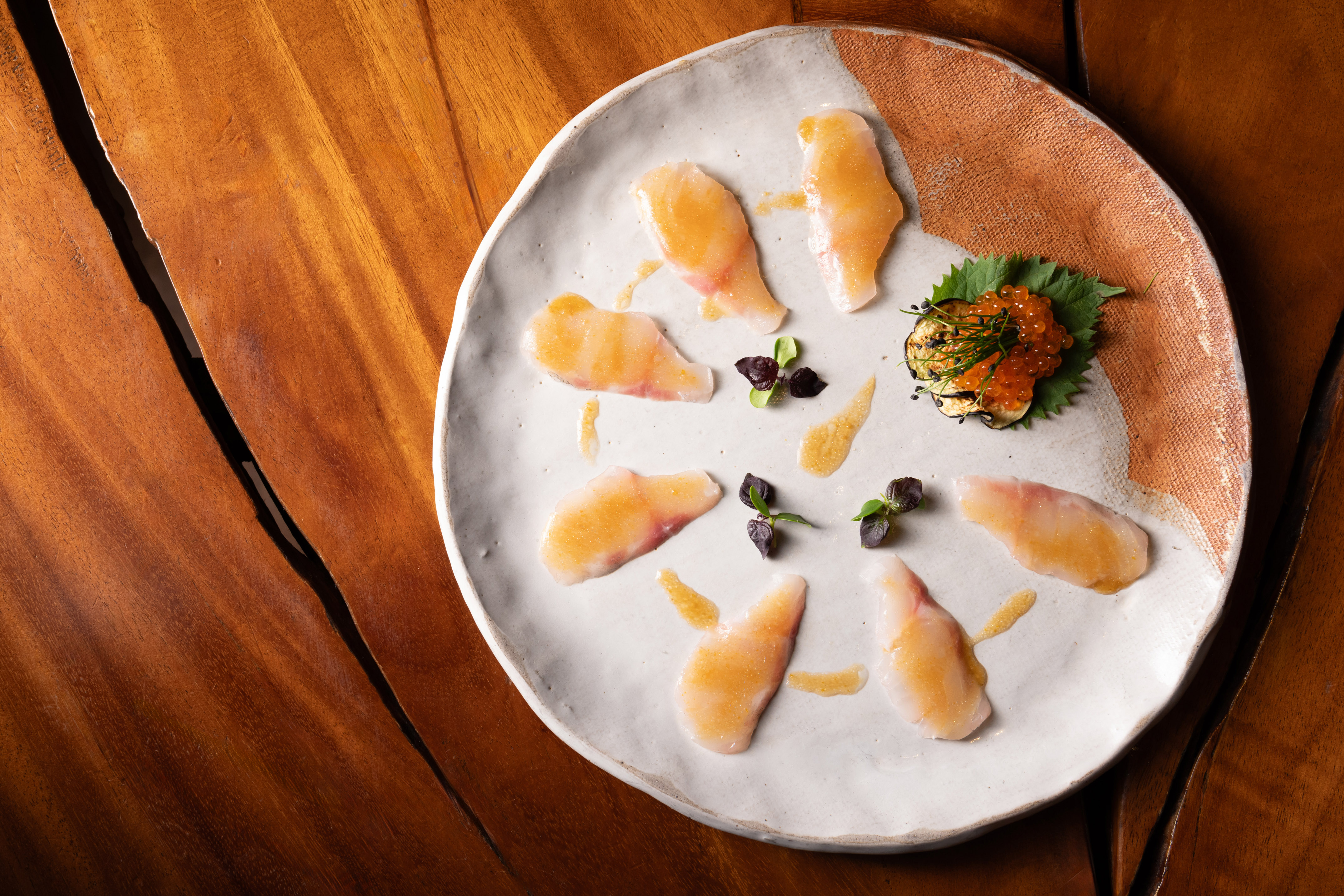 Whitefish carpaccio with ikura, microgreens, and sauce on a textured ceramic plate.