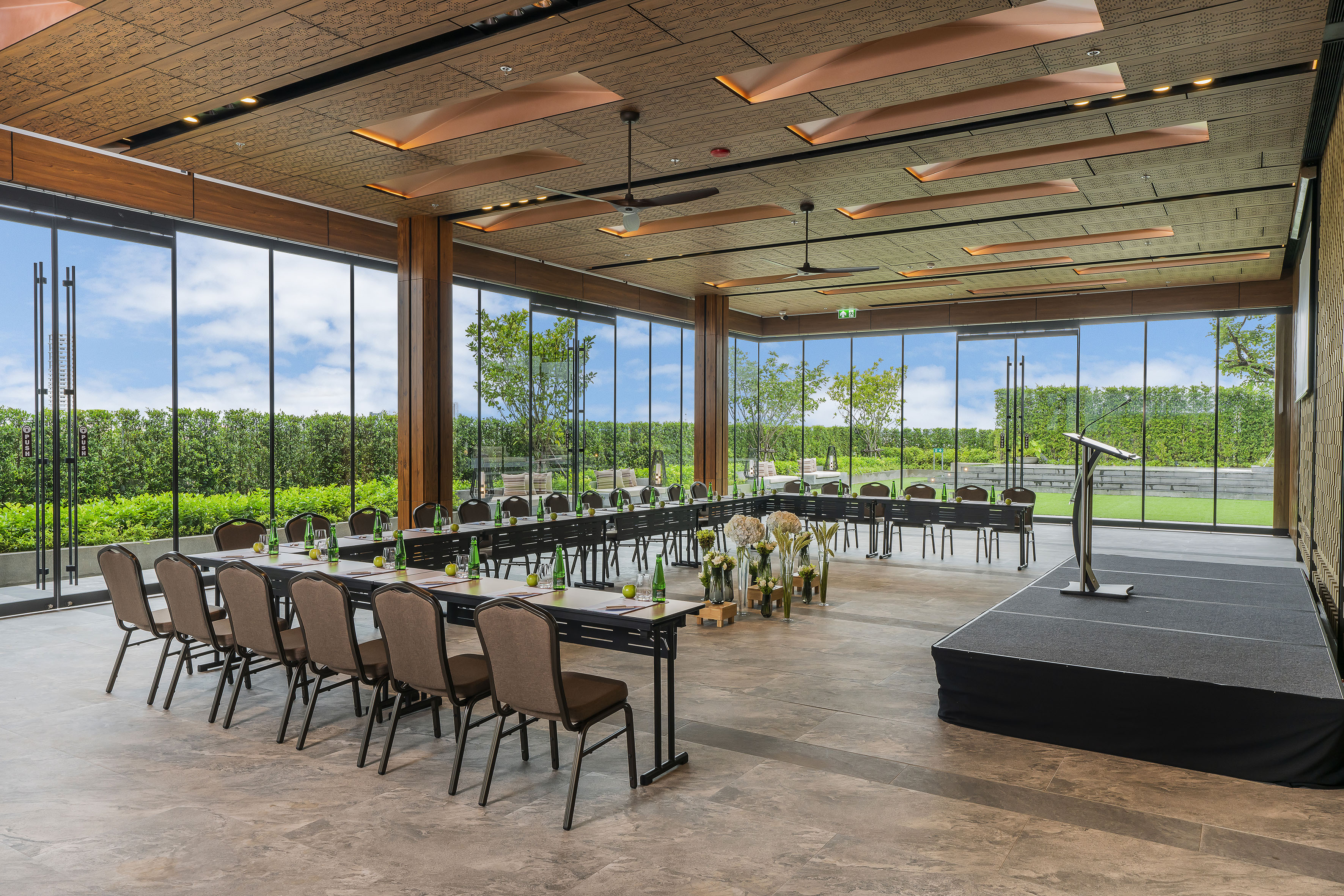 Modern conference room with U-shaped tables, stage, and garden view windows.
