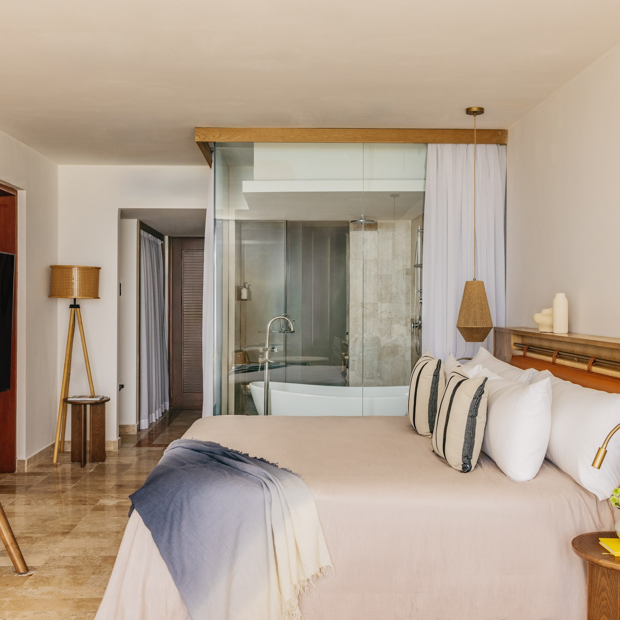 Modern hotel room with glass bathroom, bed, woven lamps, and wood accents.