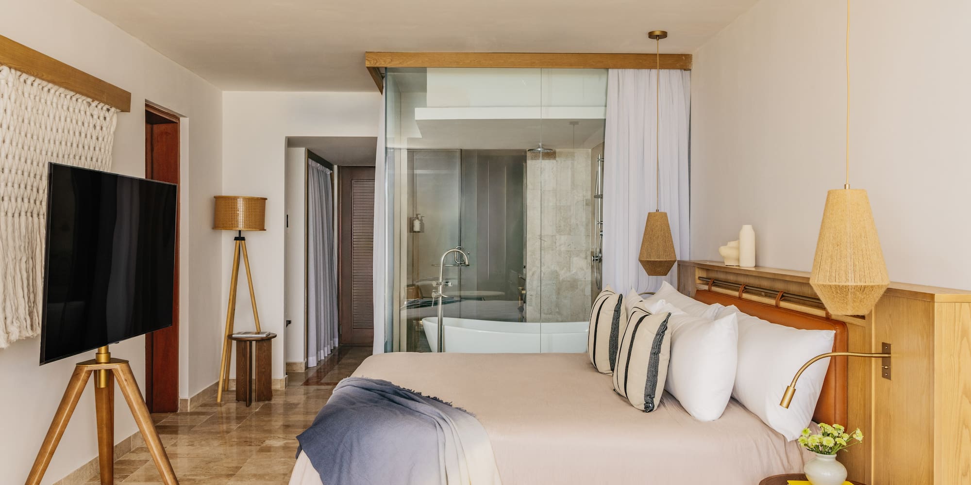 Modern hotel room with glass bathroom, bed, woven lamps, and wood accents.