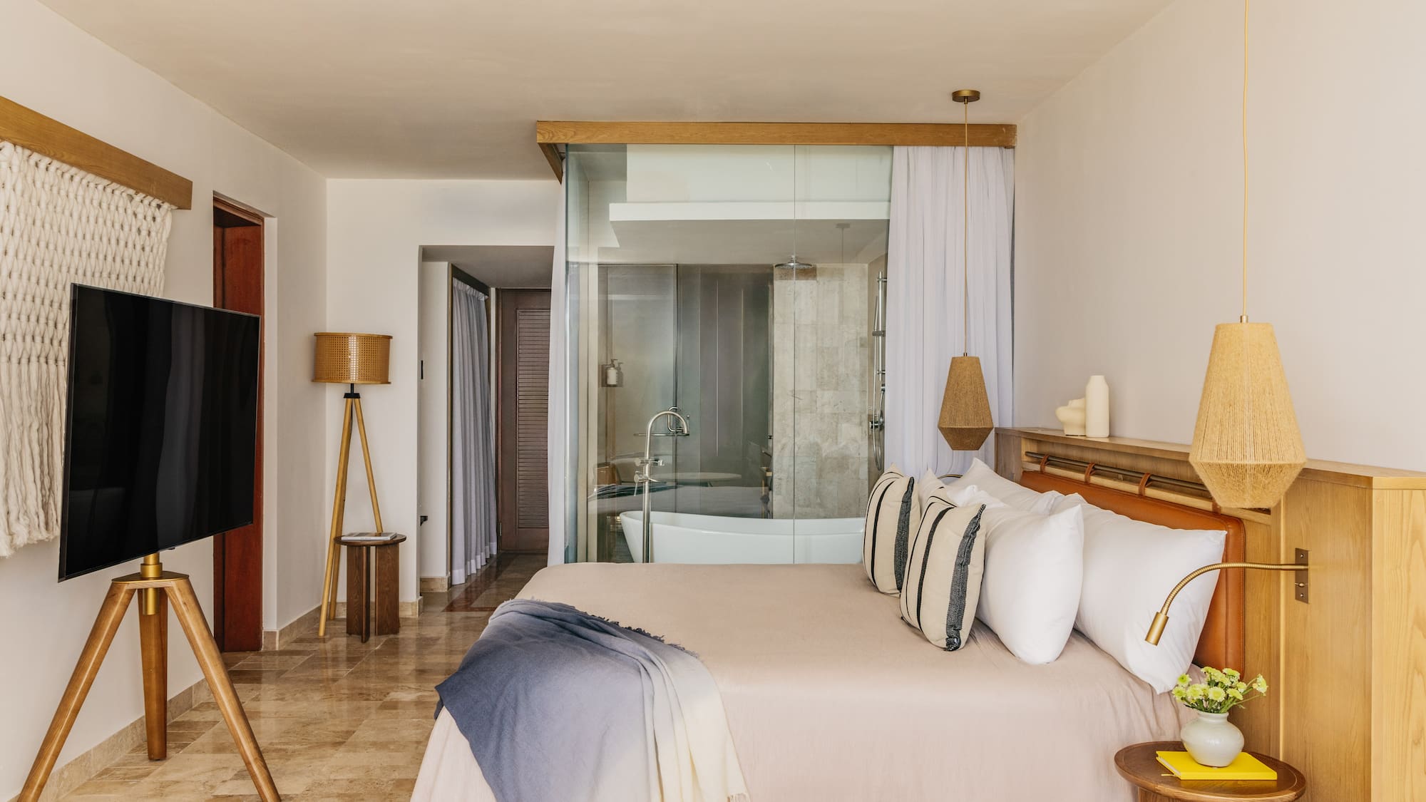 Modern hotel room with glass bathroom, bed, woven lamps, and wood accents.