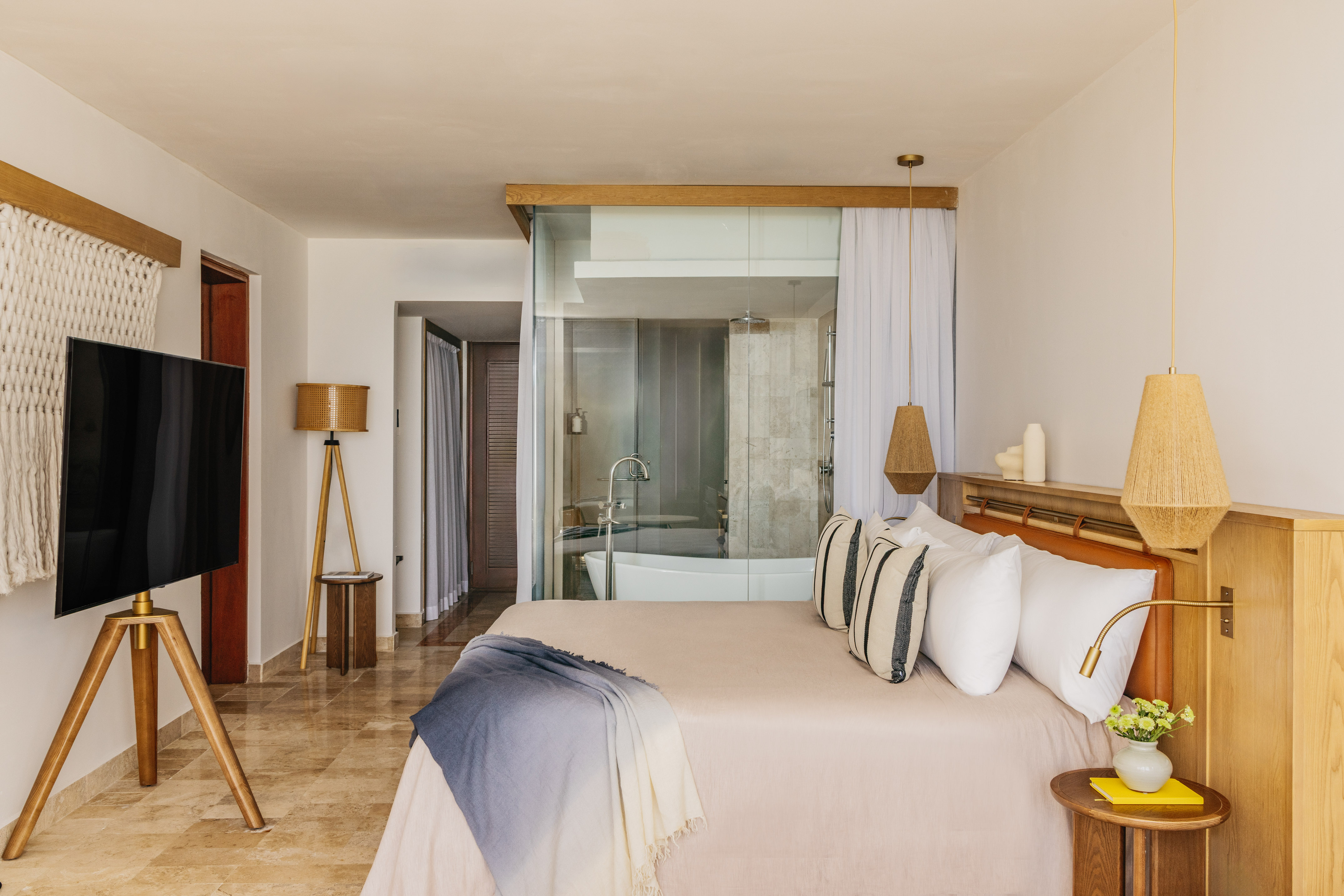 Modern hotel room with glass bathroom, bed, woven lamps, and wood accents.