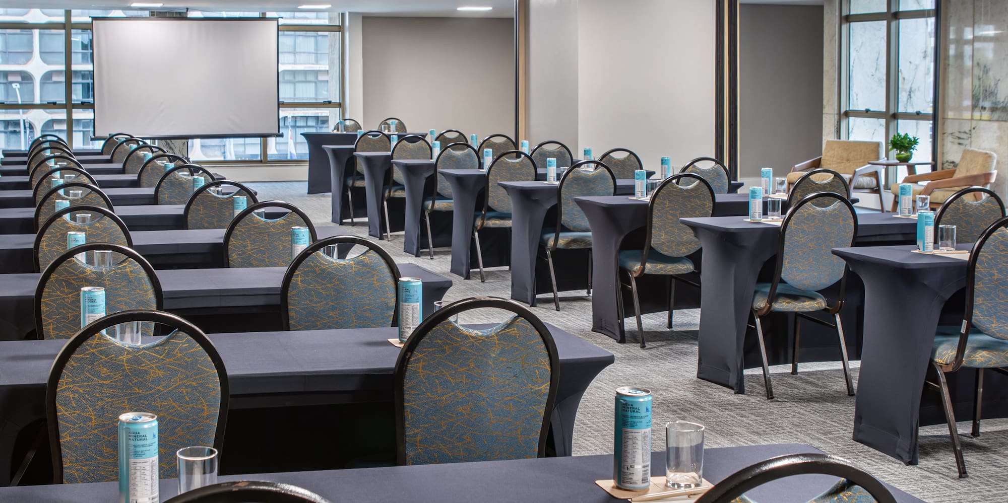 Conference room with blue chairs, dark tables, water, and a large presentation screen.