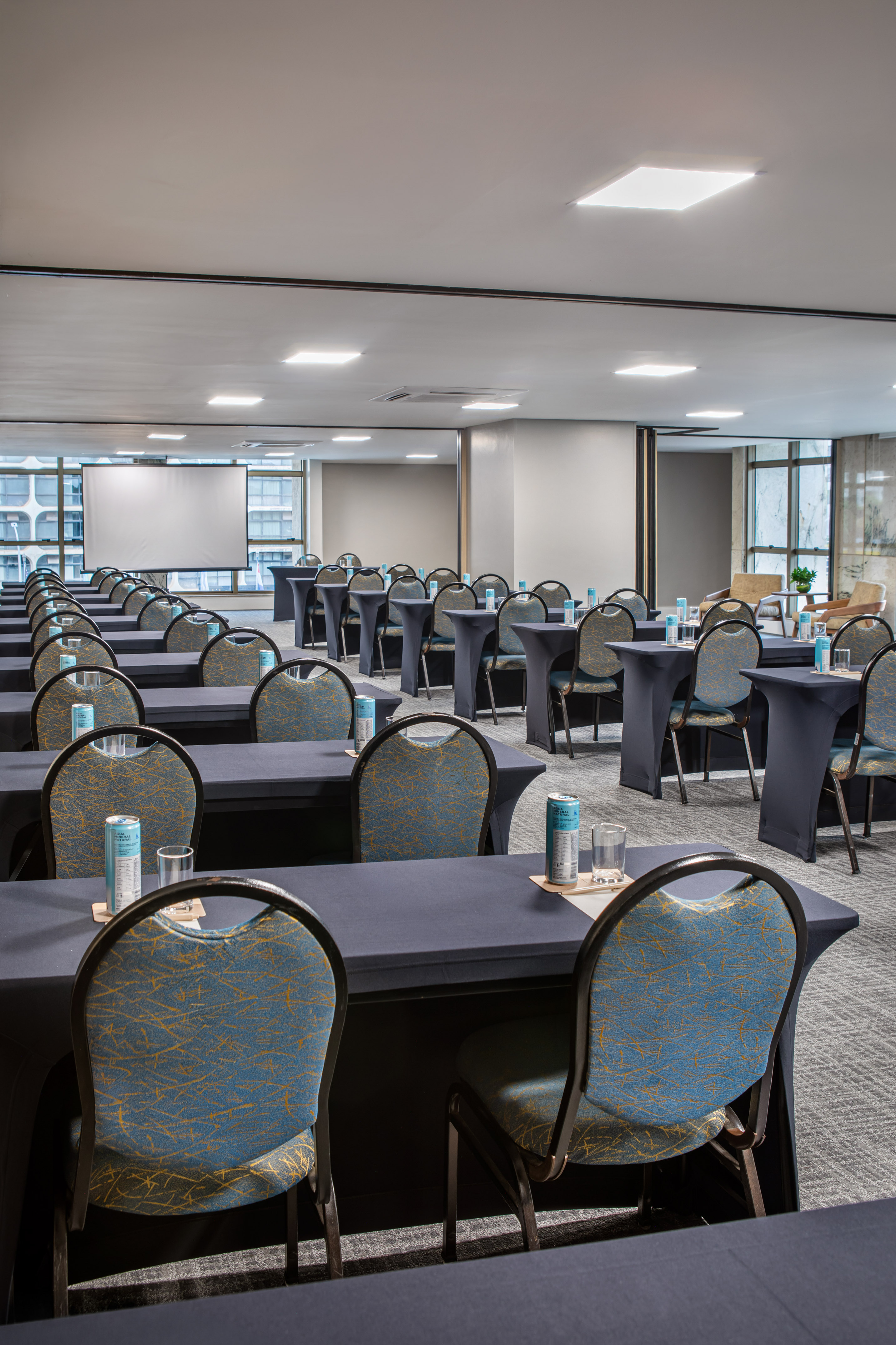 Conference room with blue chairs, dark tables, water, and a large presentation screen.