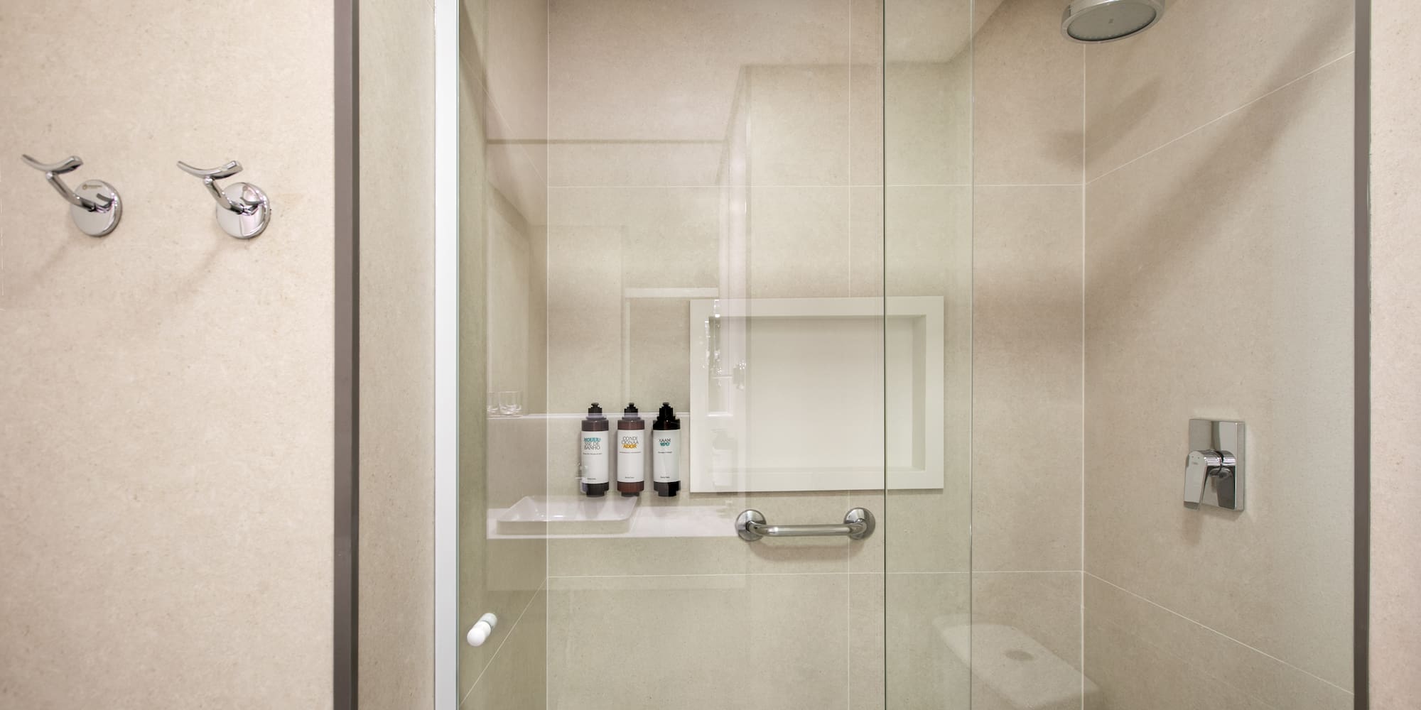 Modern glass shower with neutral tiled walls, toiletries, grab bar, and hooks.