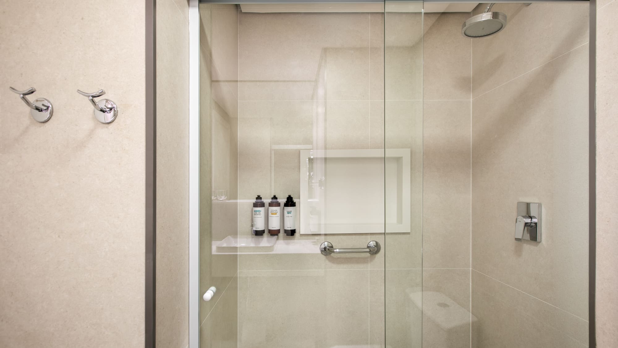 Modern glass shower with neutral tiled walls, toiletries, grab bar, and hooks.