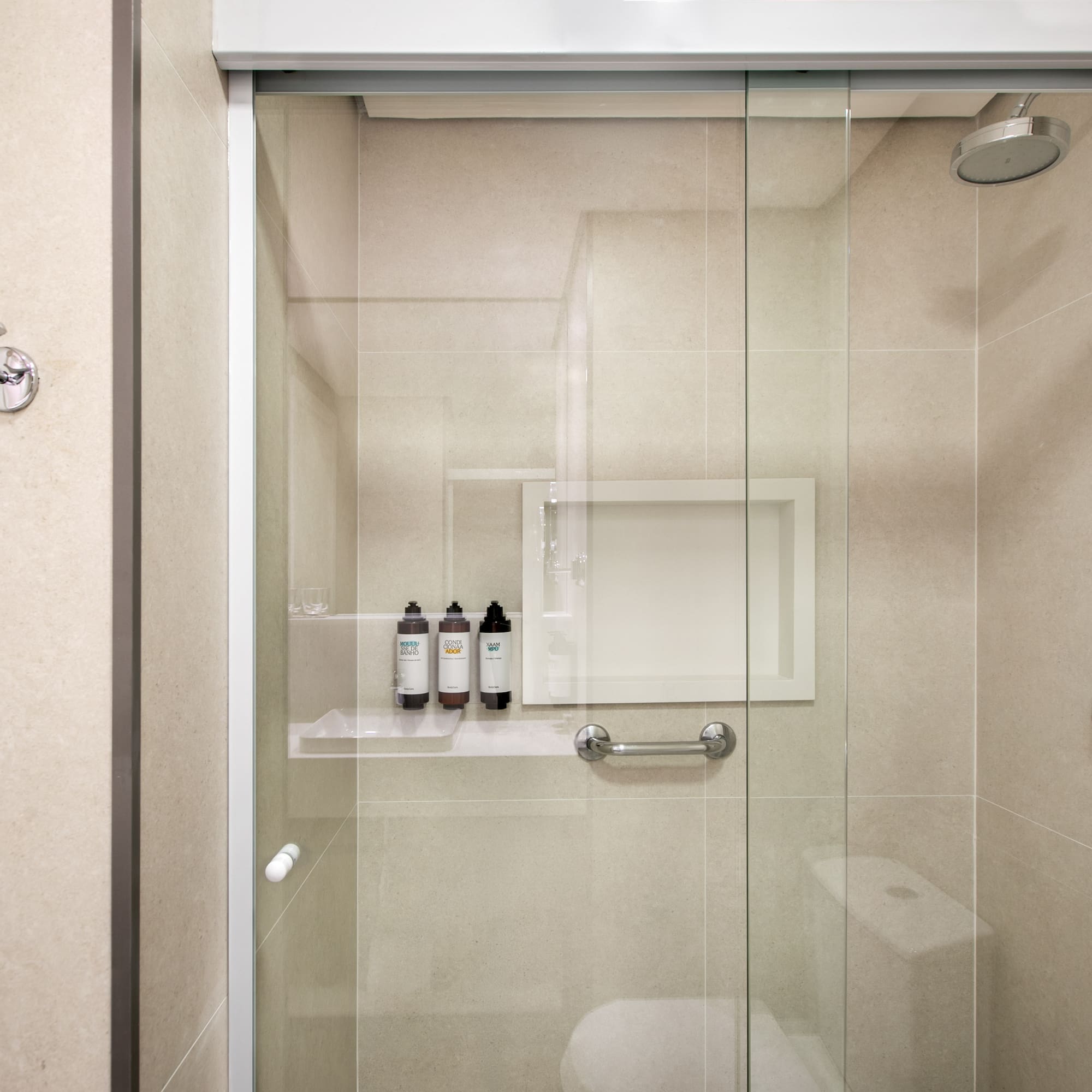 Modern glass shower with neutral tiled walls, toiletries, grab bar, and hooks.