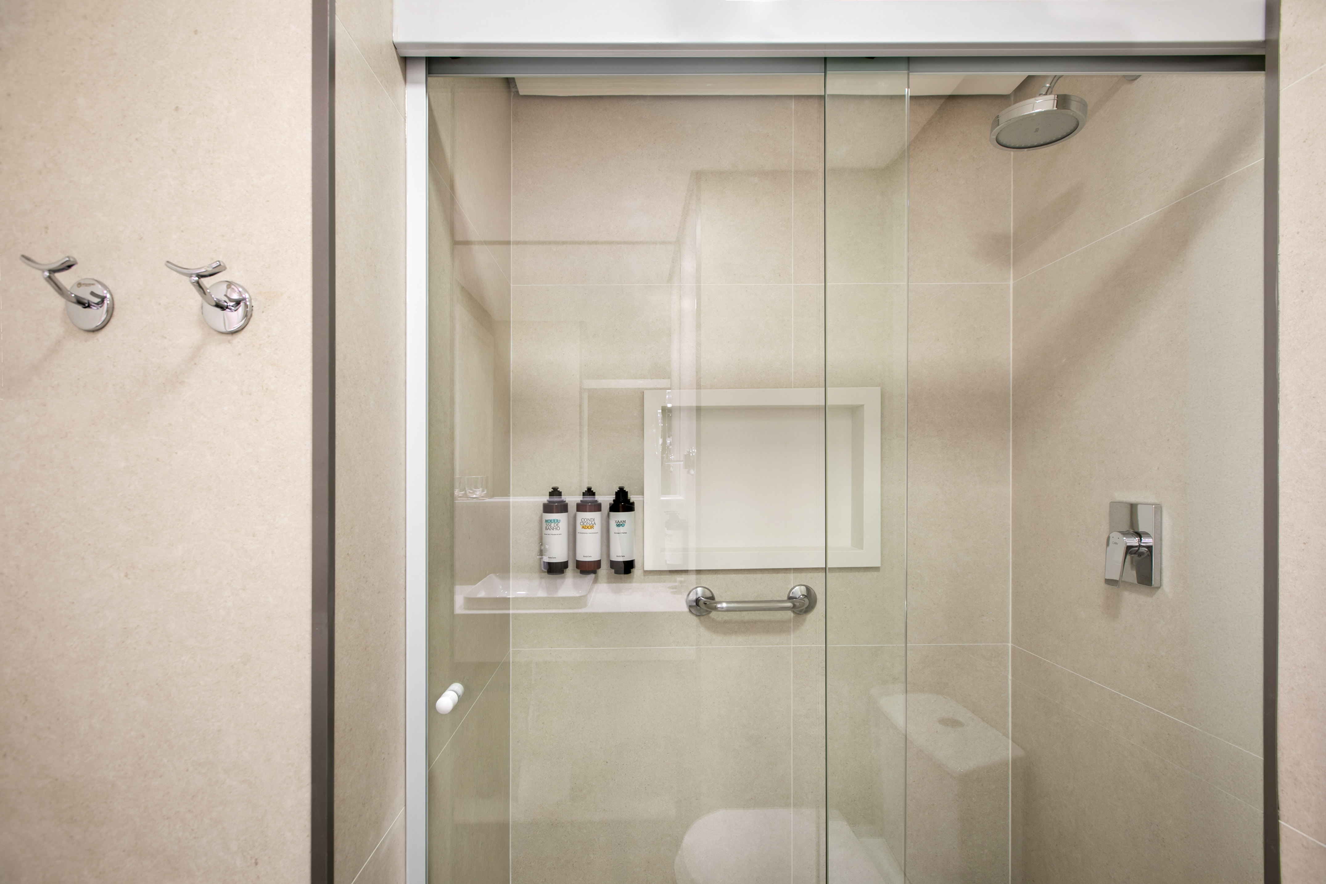 Modern glass shower with neutral tiled walls, toiletries, grab bar, and hooks.