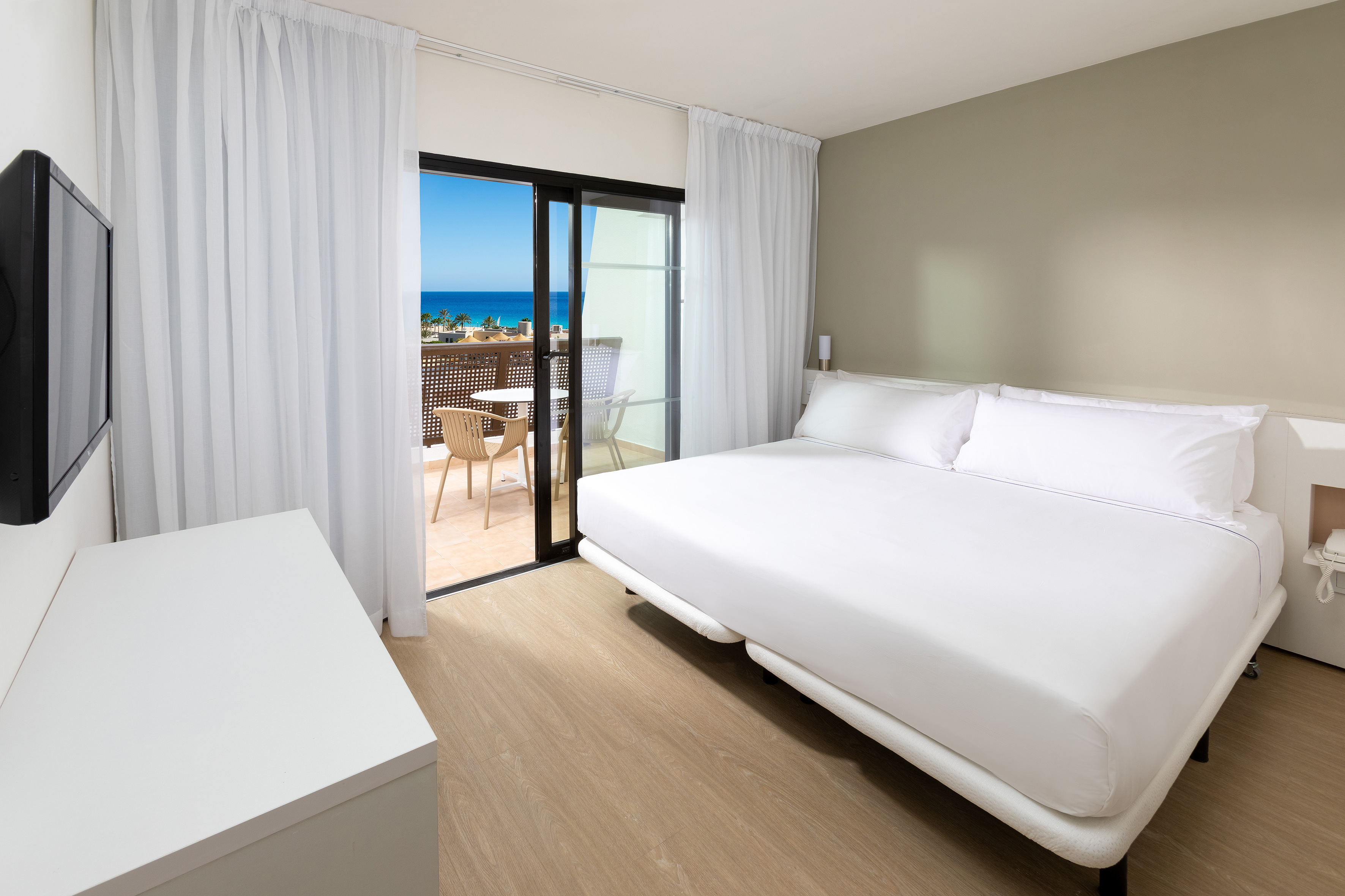 Modern hotel bedroom with white bed and balcony overlooking the ocean.