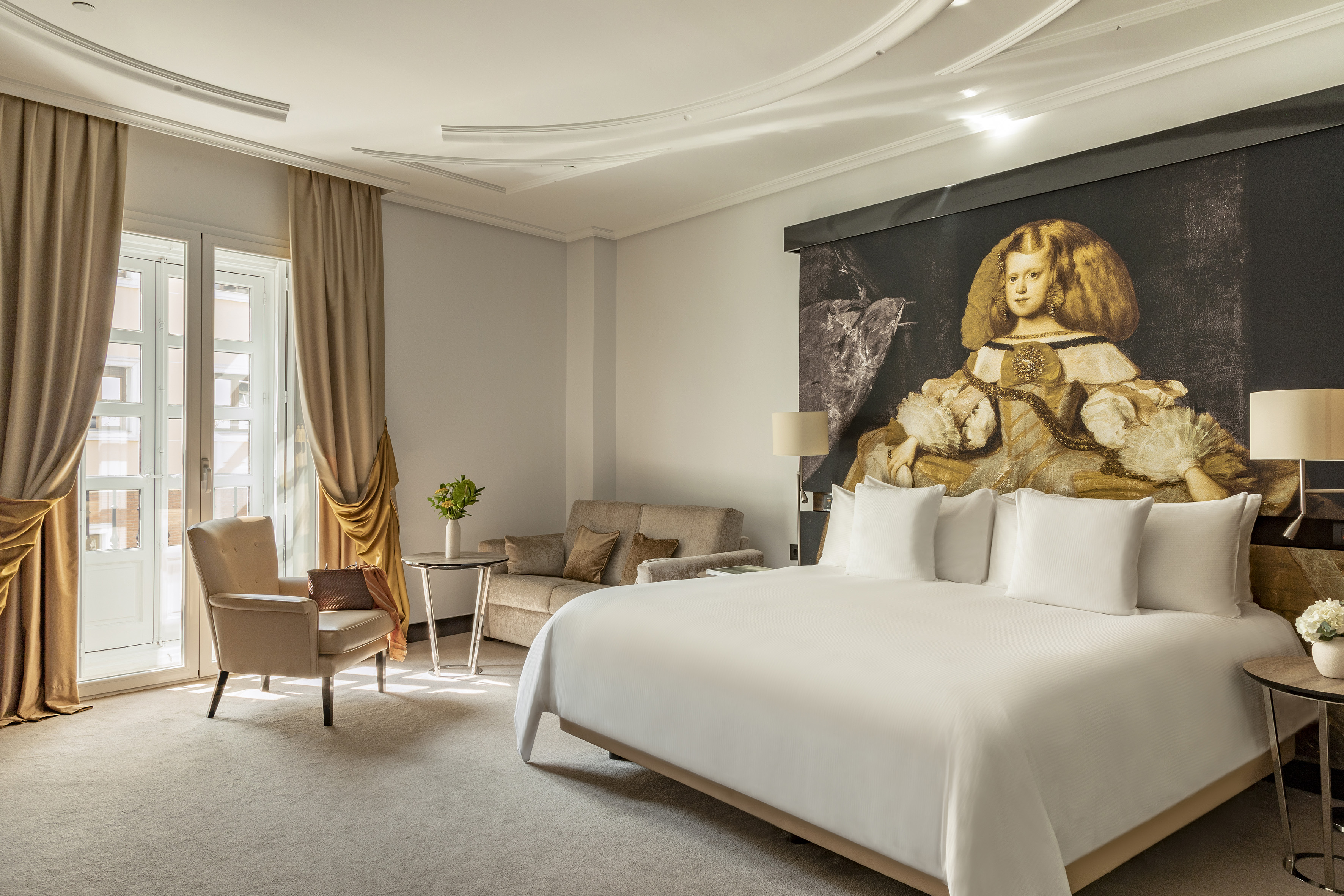 Modern hotel suite, king bed, large Velázquez artwork, armchair, and balcony.