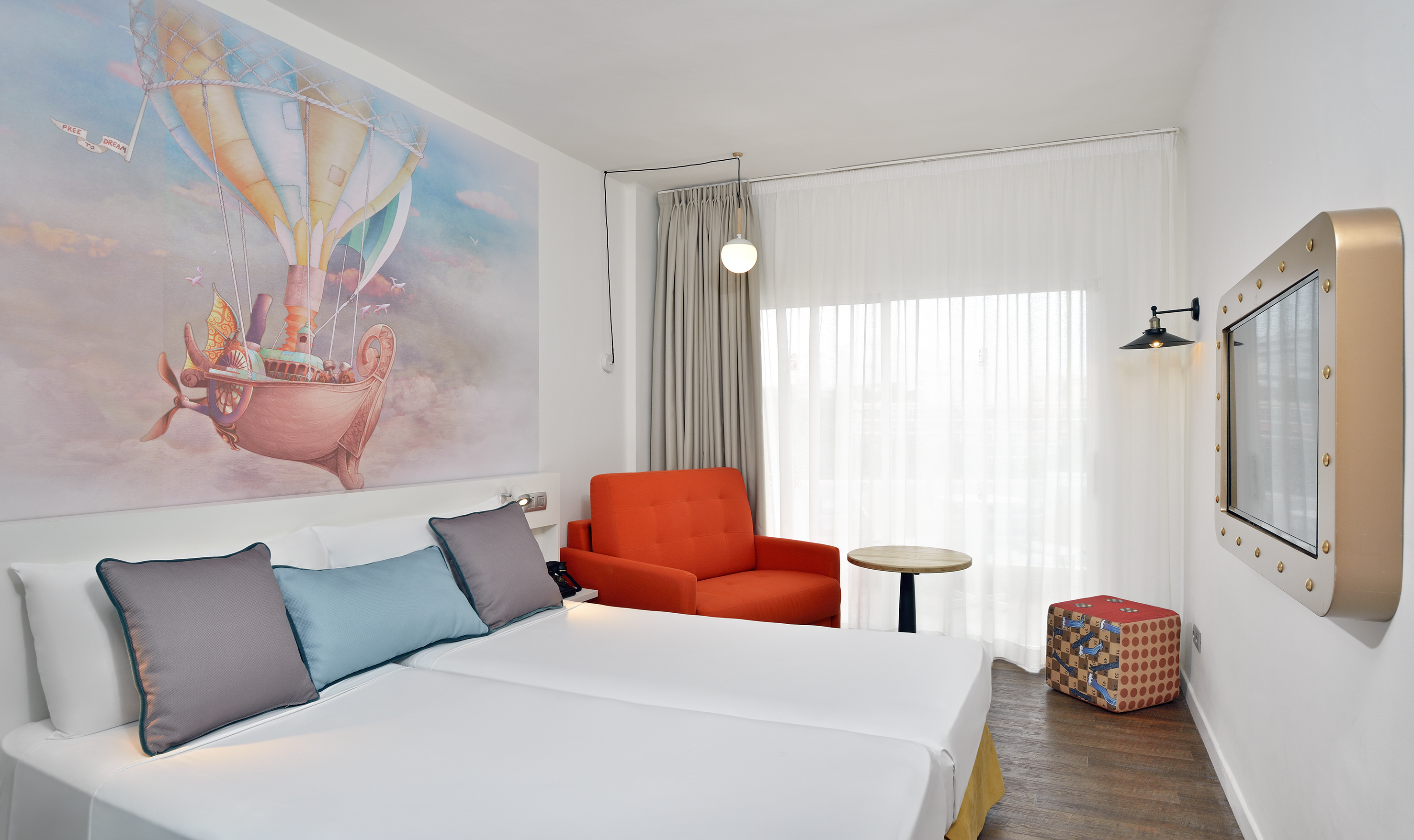 Bright hotel room with hot air balloon mural, twin beds, orange armchair, TV.