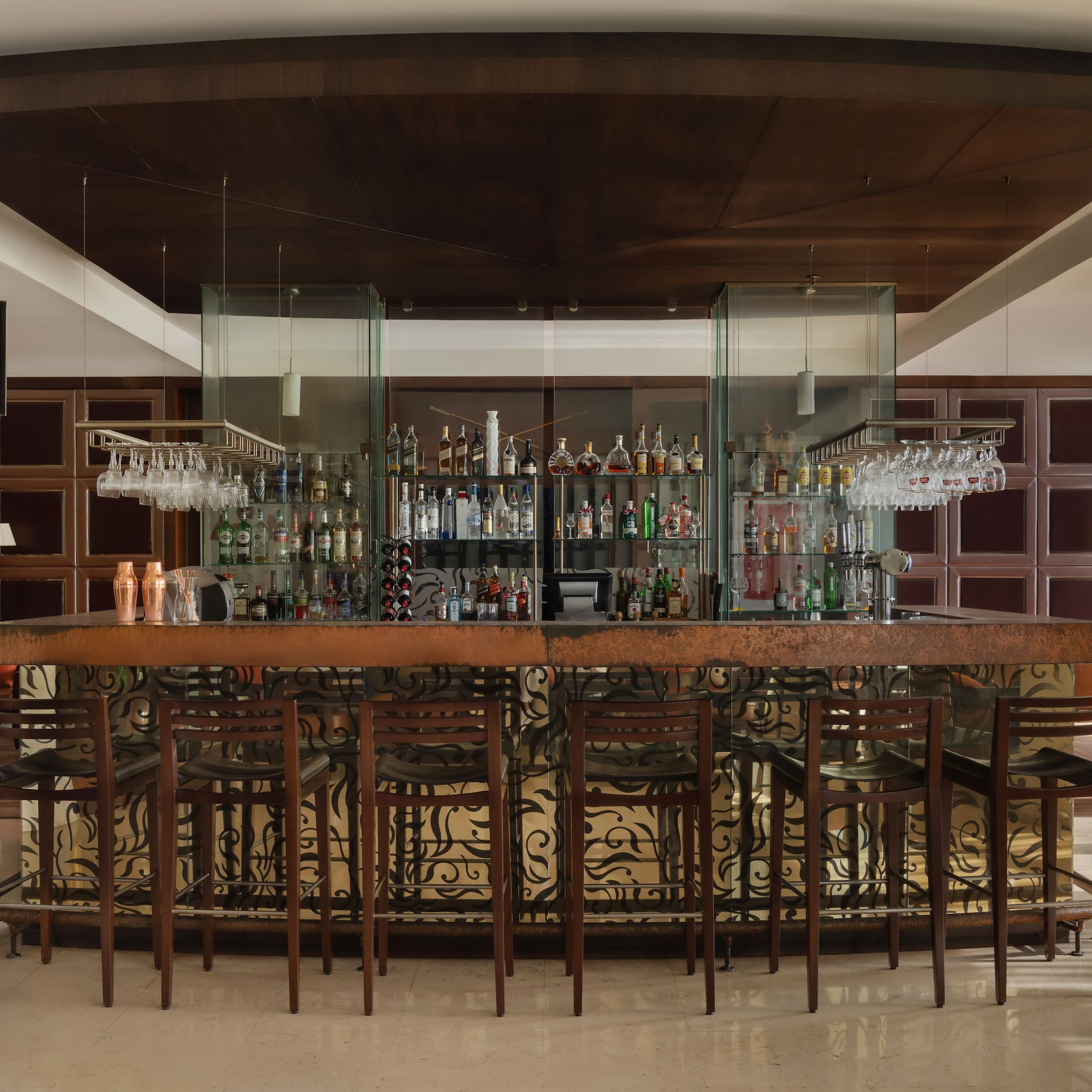 Hotel bar with stocked liquor bottles, dark wood stools, and decorative ceiling.