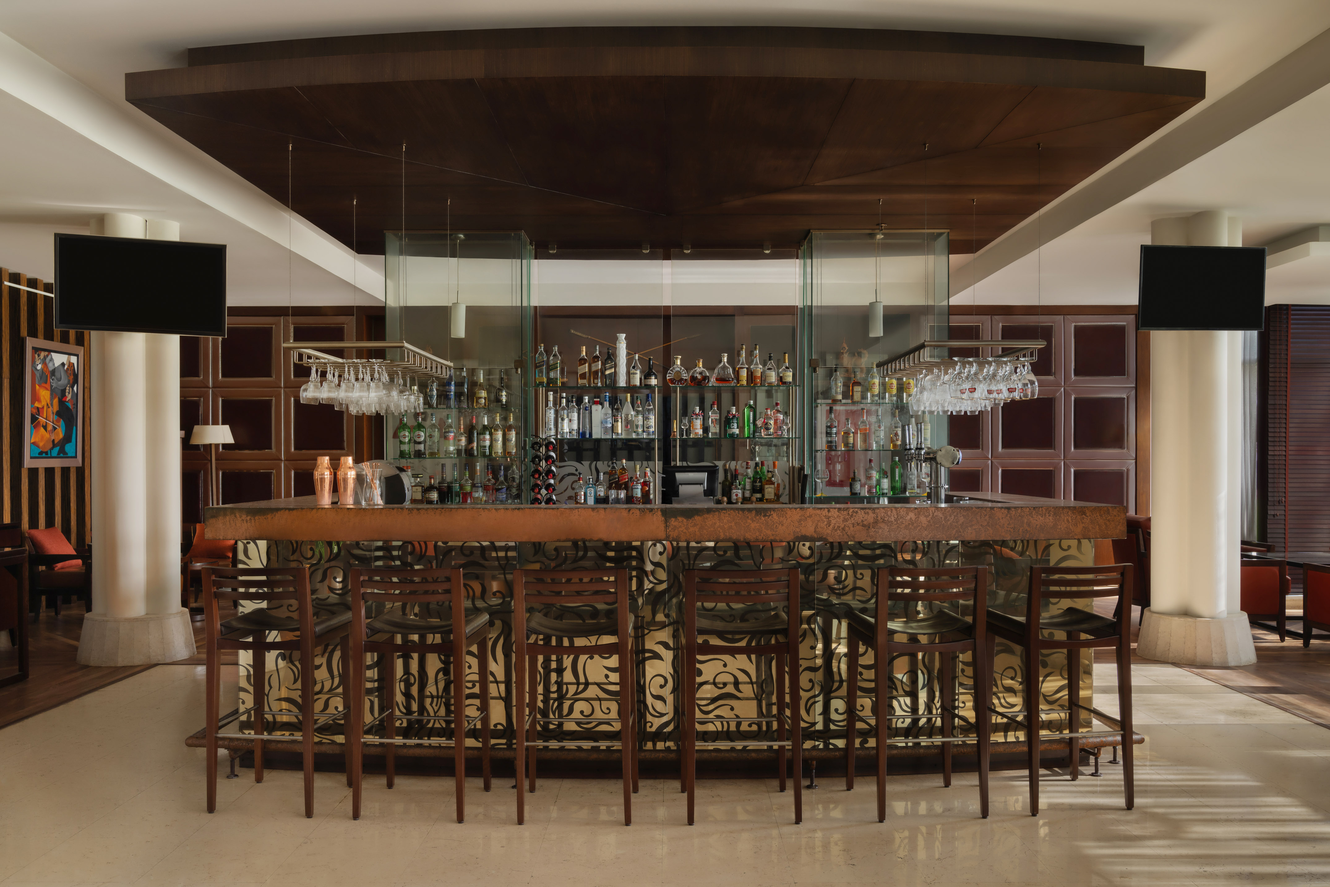 Hotel bar with stocked liquor bottles, dark wood stools, and decorative ceiling.