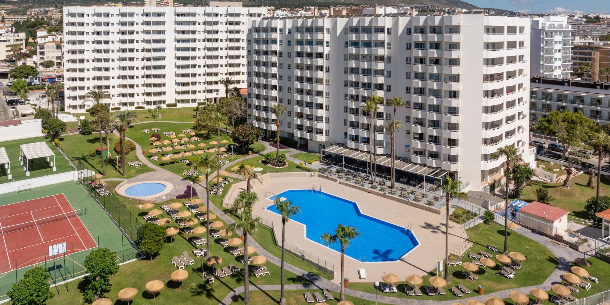 Hotel resort with pools, tennis court, palm trees, and white apartment buildings.