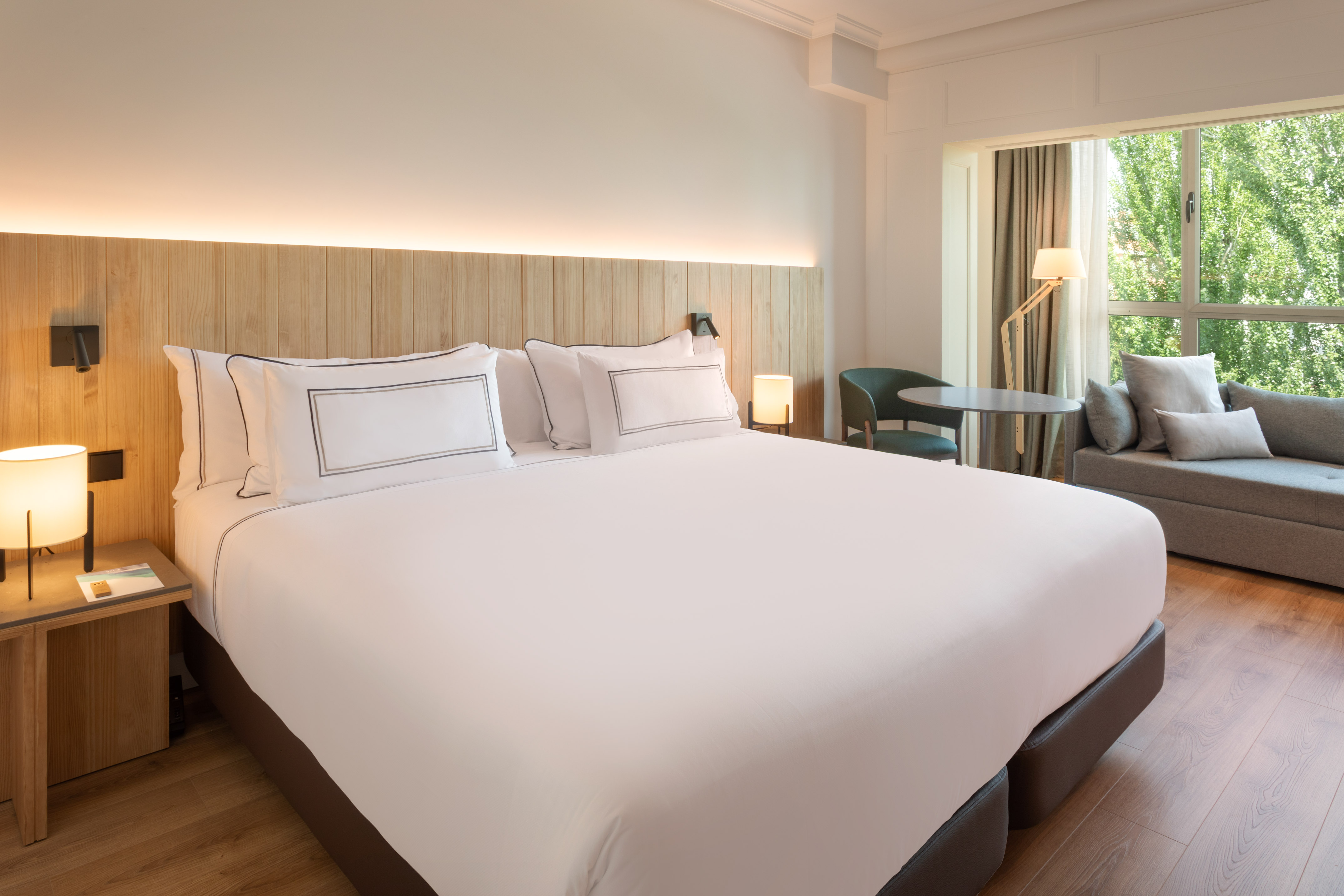 Modern hotel room with white bed, wooden headboard, and bright window seating.