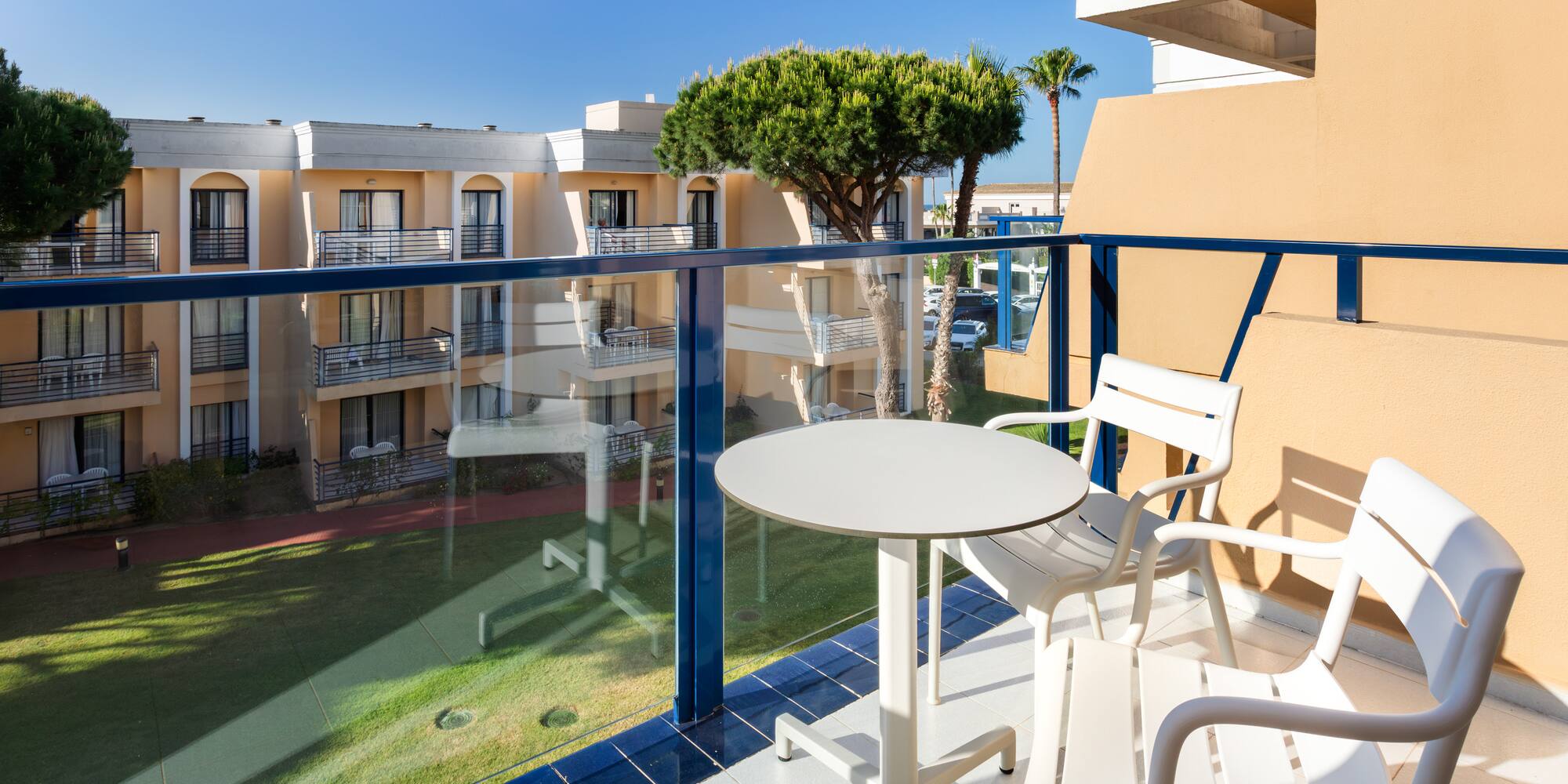 Sunny hotel balcony with white table, chairs, resort view, lush lawn, blue sky.