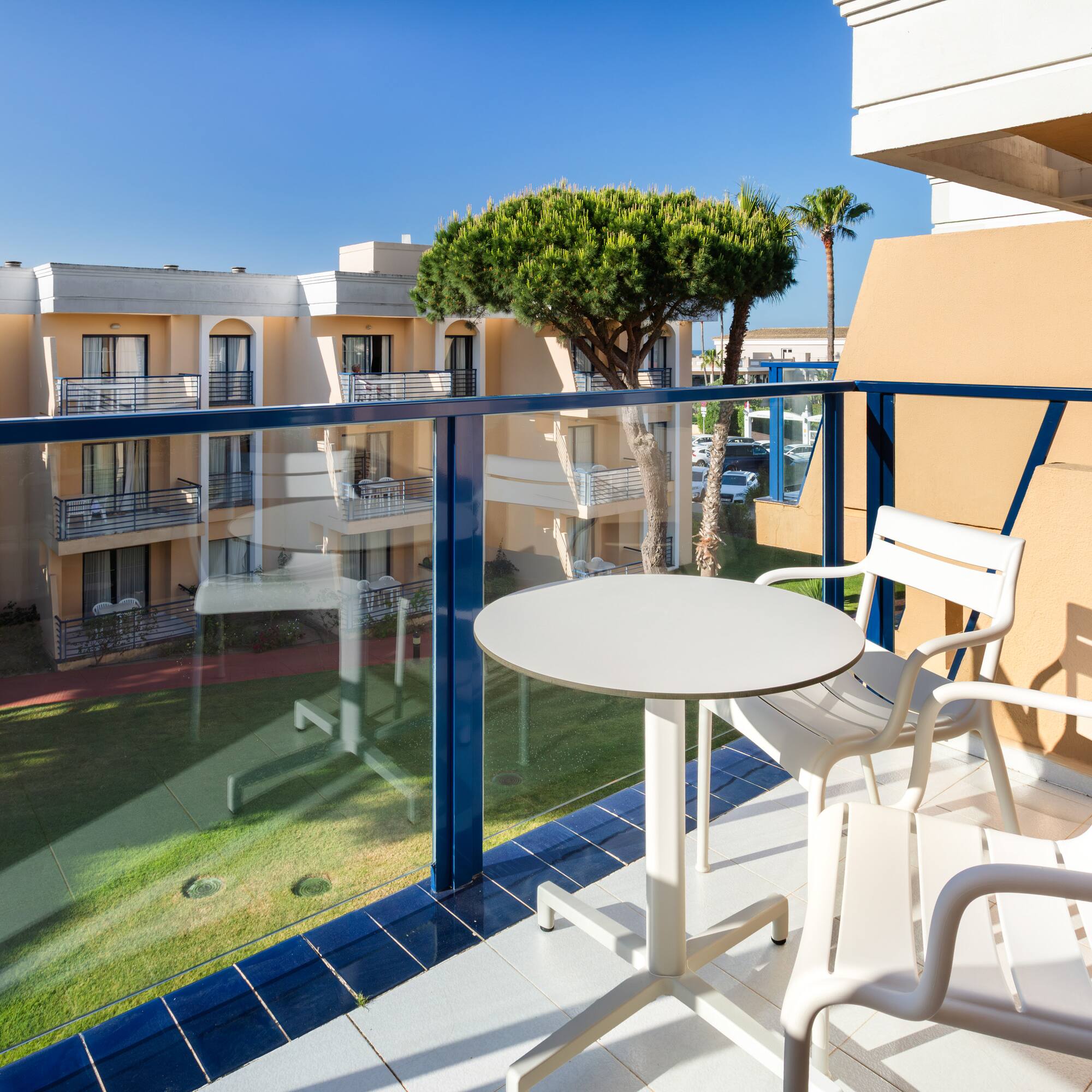 Sunny hotel balcony with white table, chairs, resort view, lush lawn, blue sky.