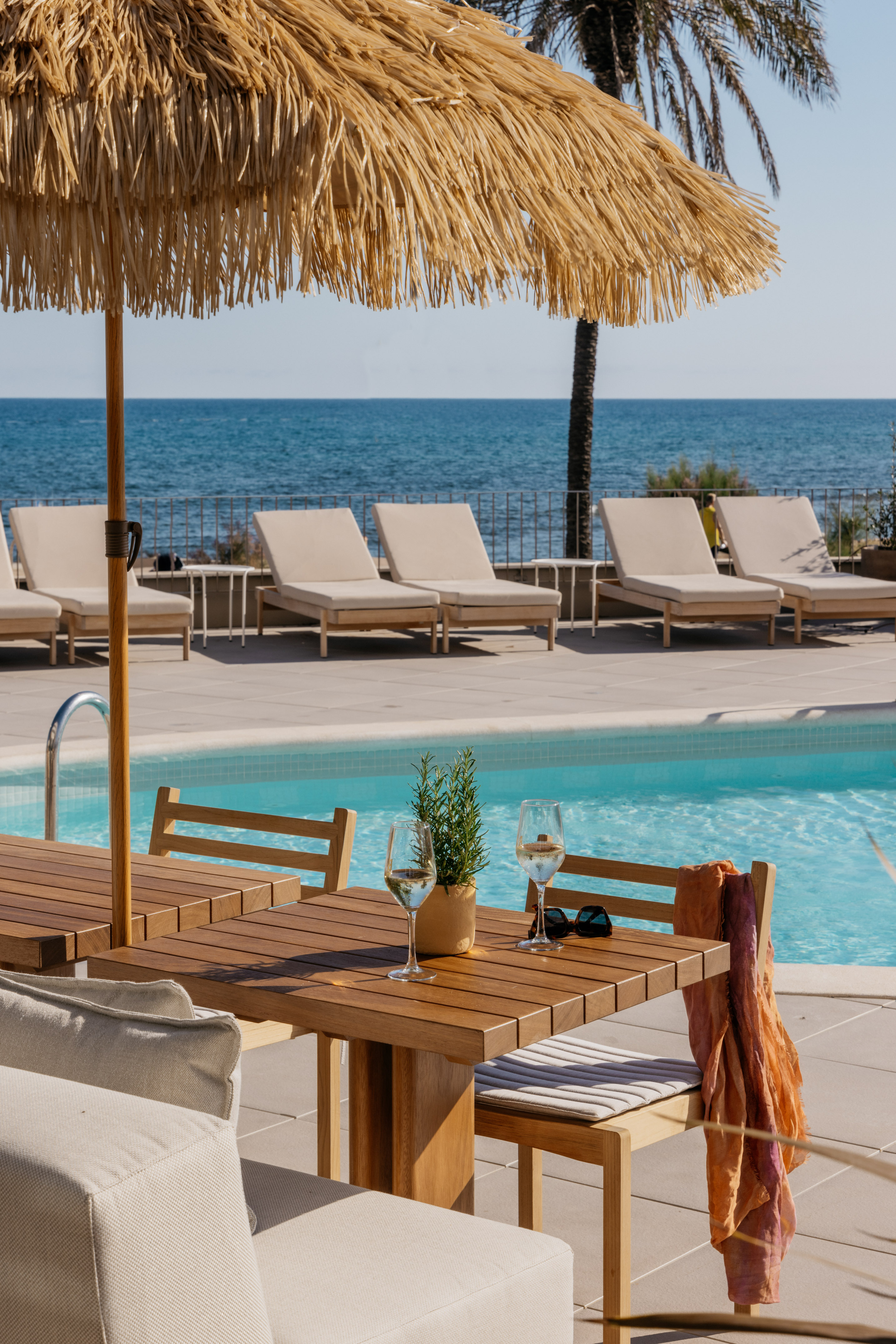 Resort poolside with straw umbrella, wooden table, wine, and ocean view.