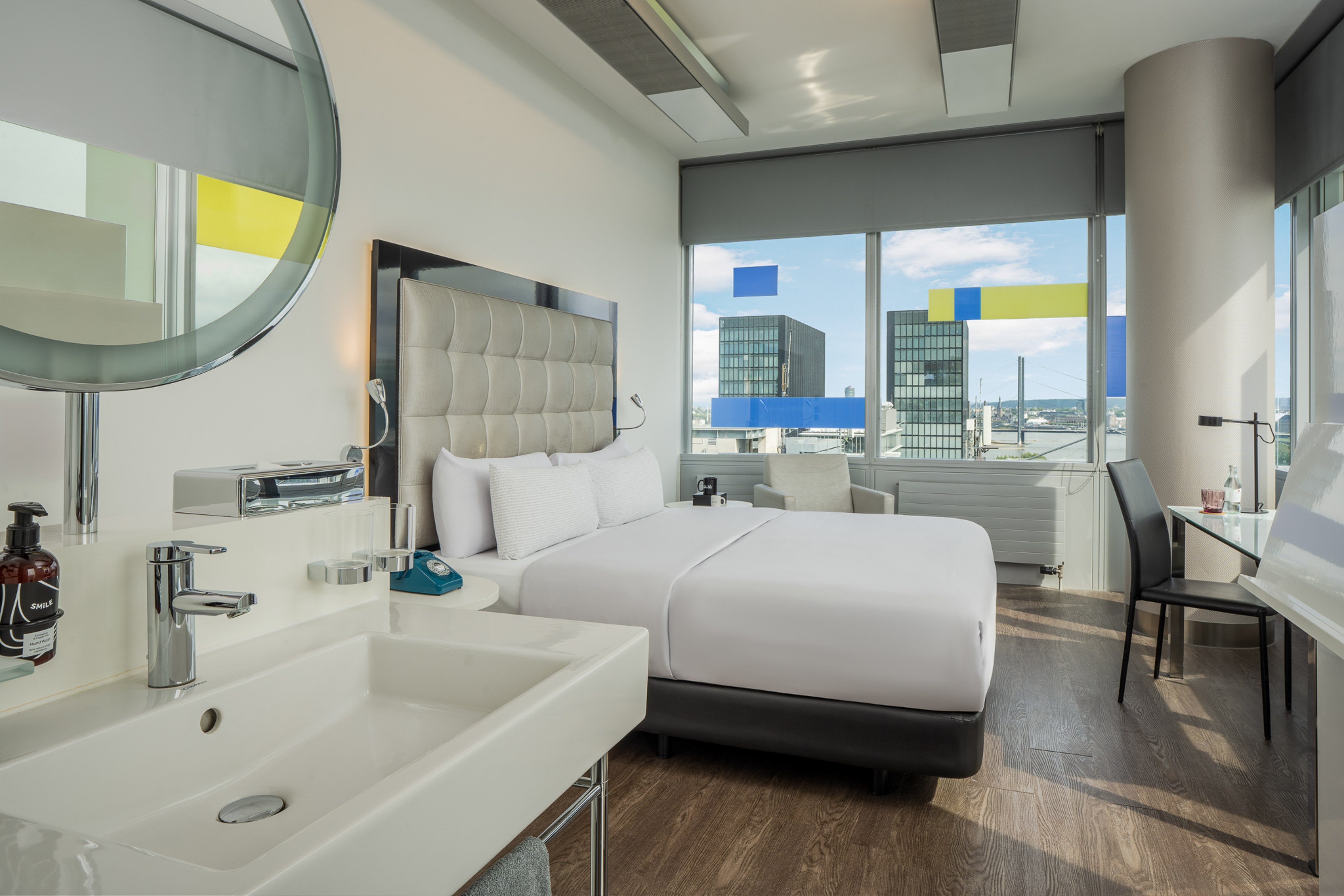 Modern luxury hotel room with bed, sink, desk, and city view.