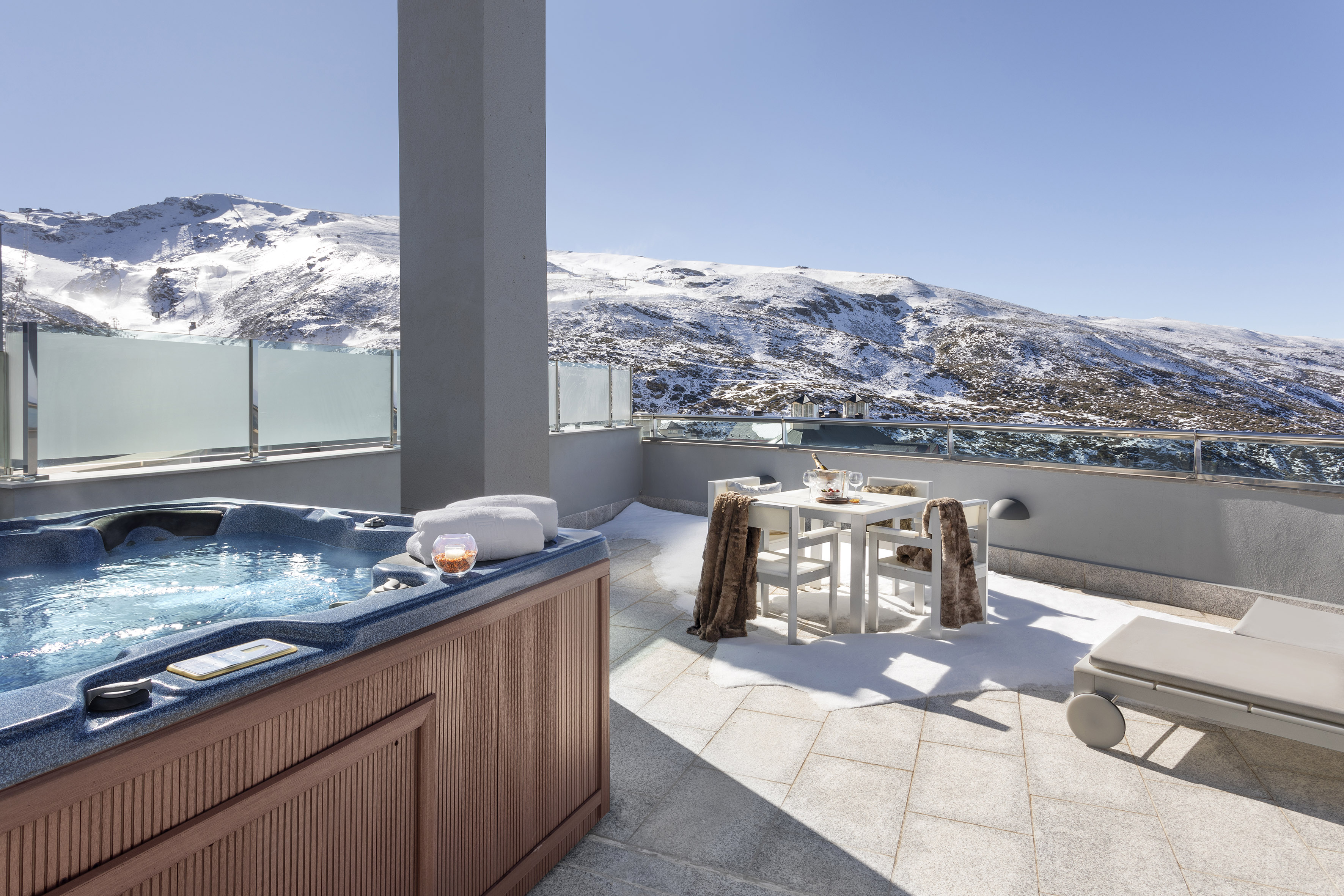Outdoor hot tub on snowy balcony with mountain view and dining table.