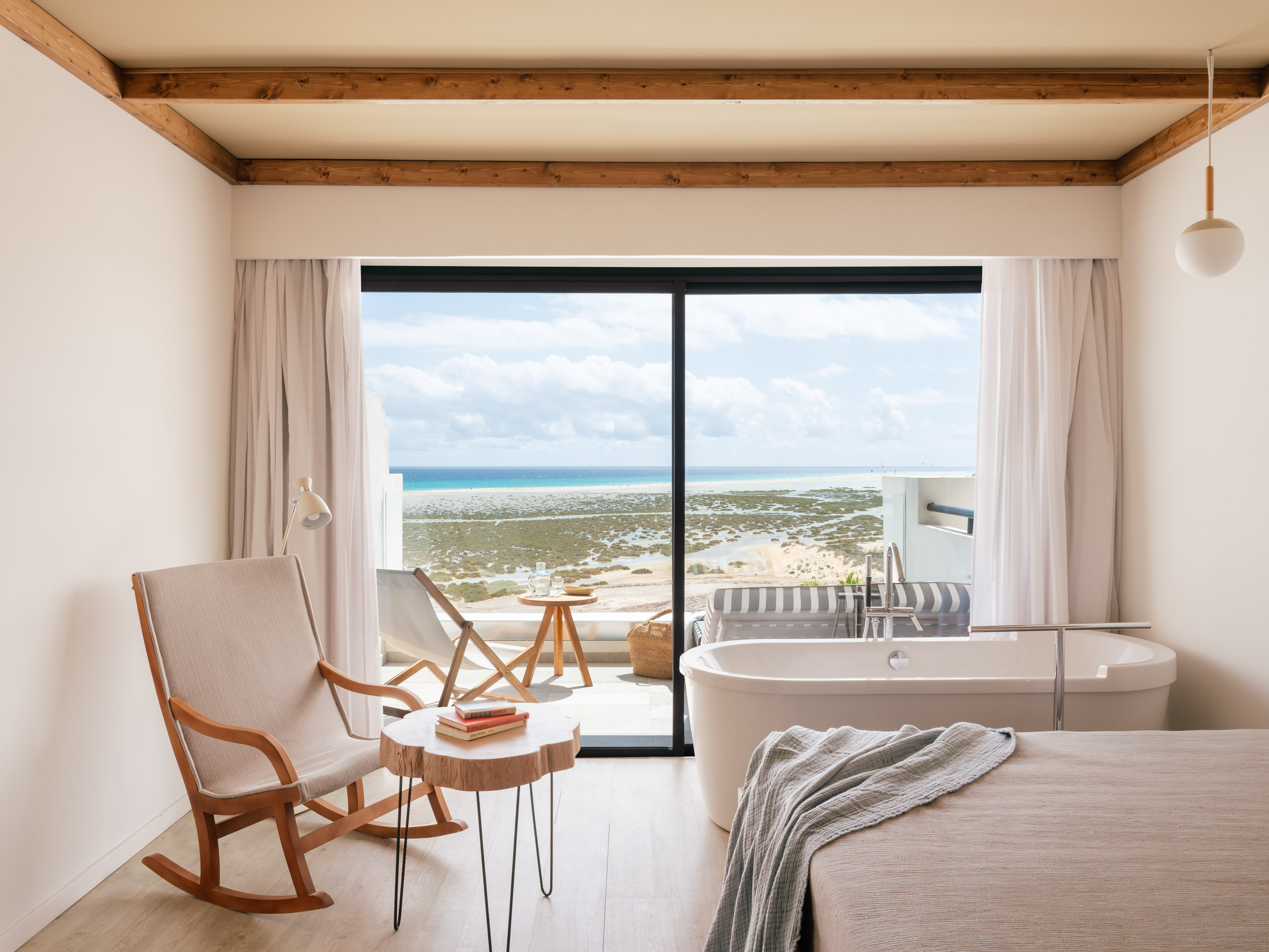 Habitación moderna con vistas al mar, bañera junto al ventanal y mecedora de madera.