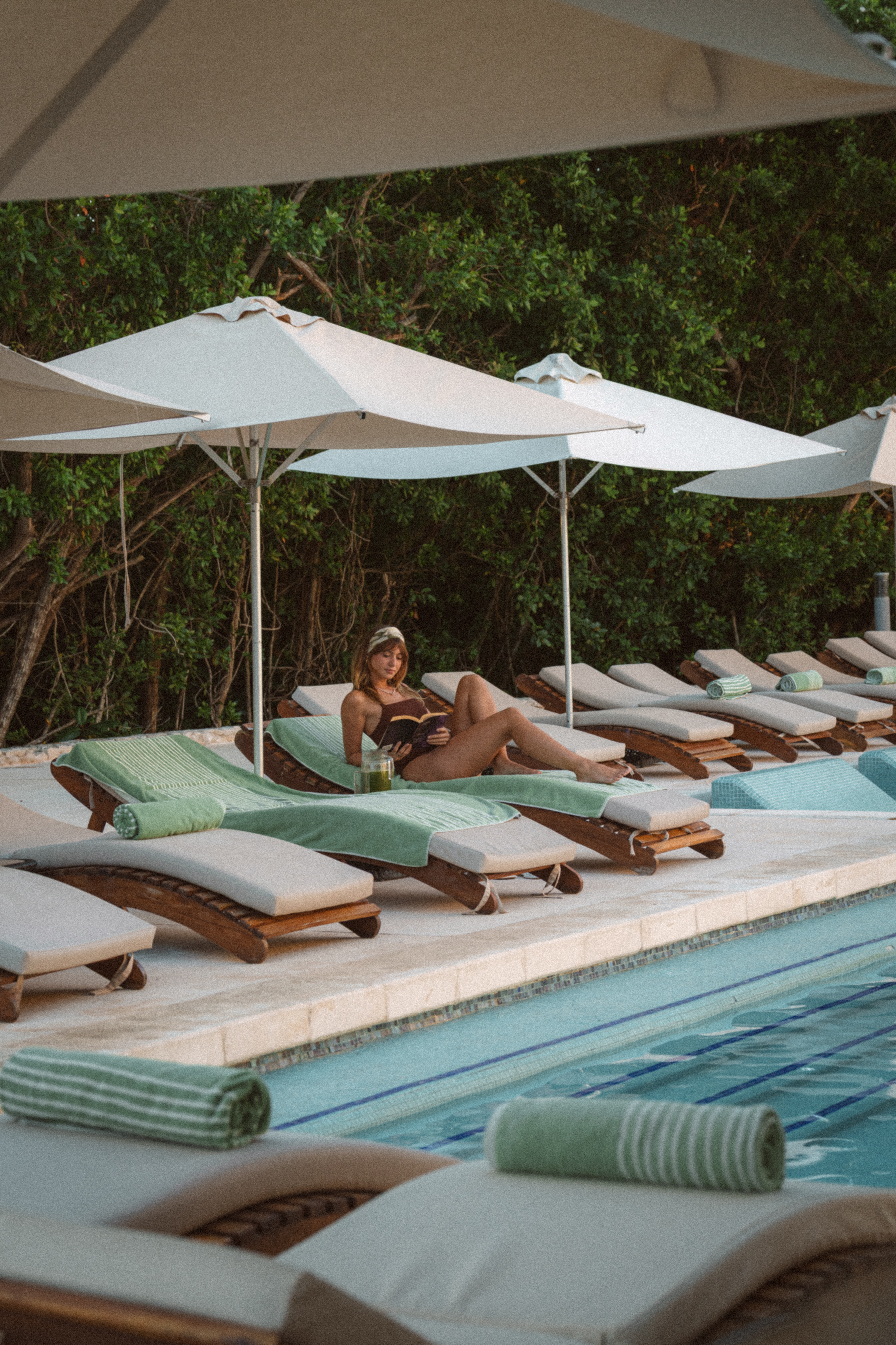 Woman reading on poolside lounge chair under umbrella at a resort.