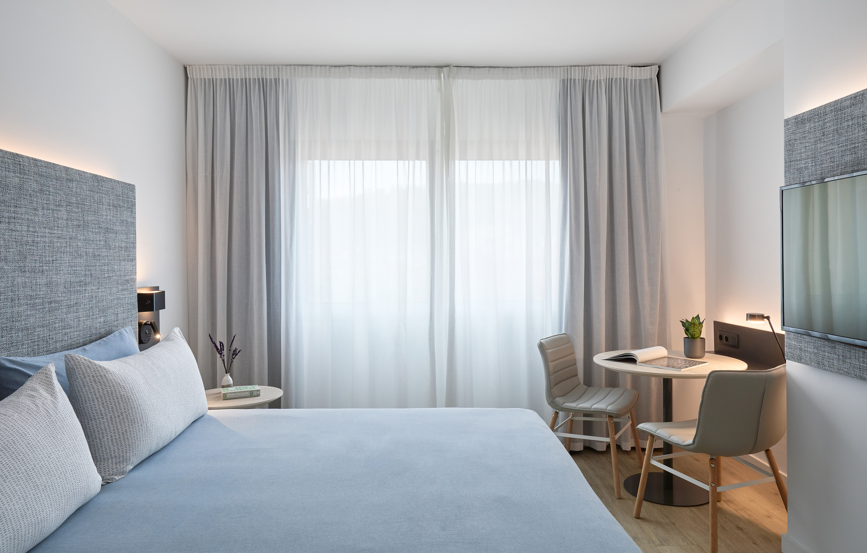 Modern hotel bedroom with light blue bed, grey headboard, window, and small table.