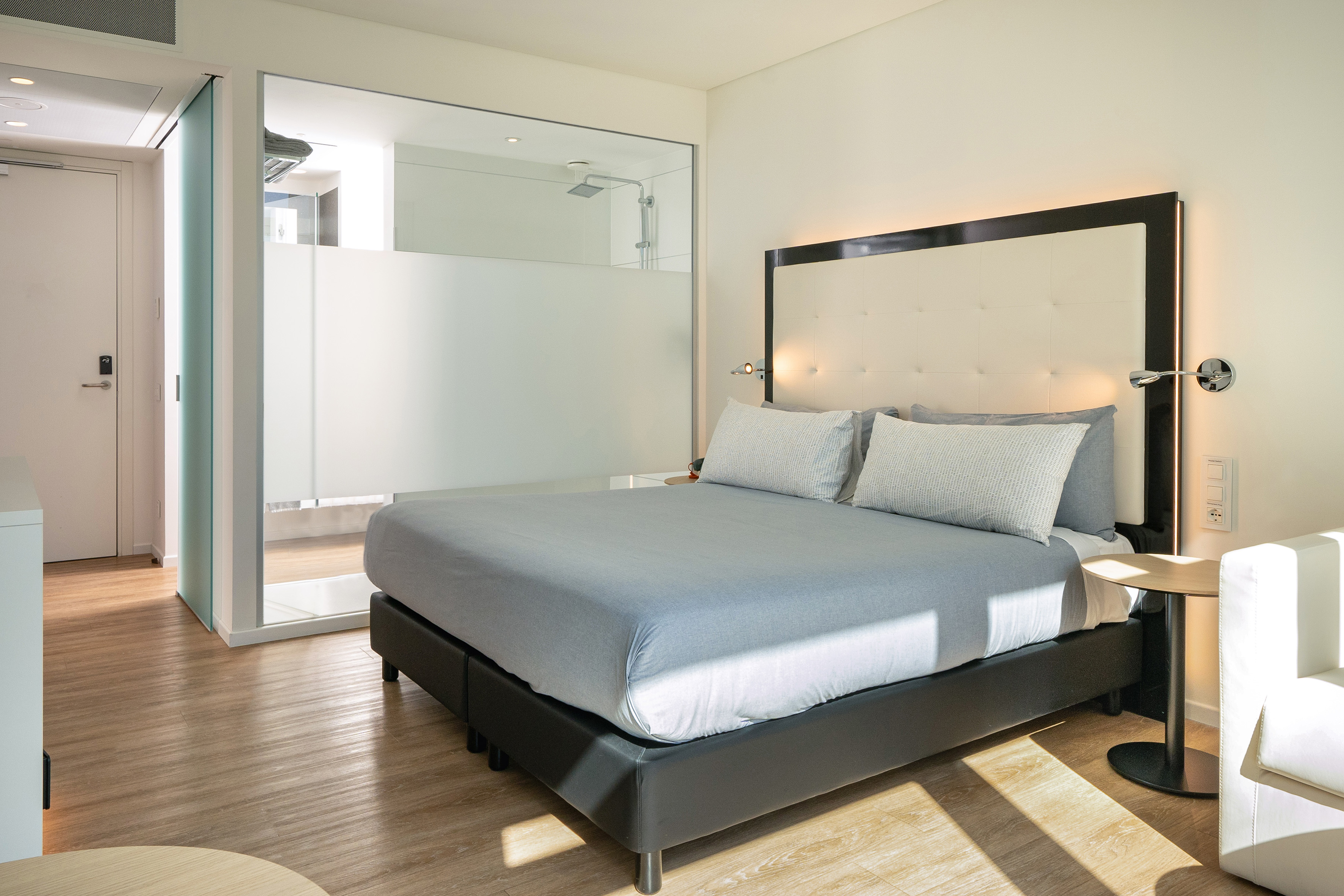 Modern hotel bedroom with queen bed, grey bedding, and glass shower.