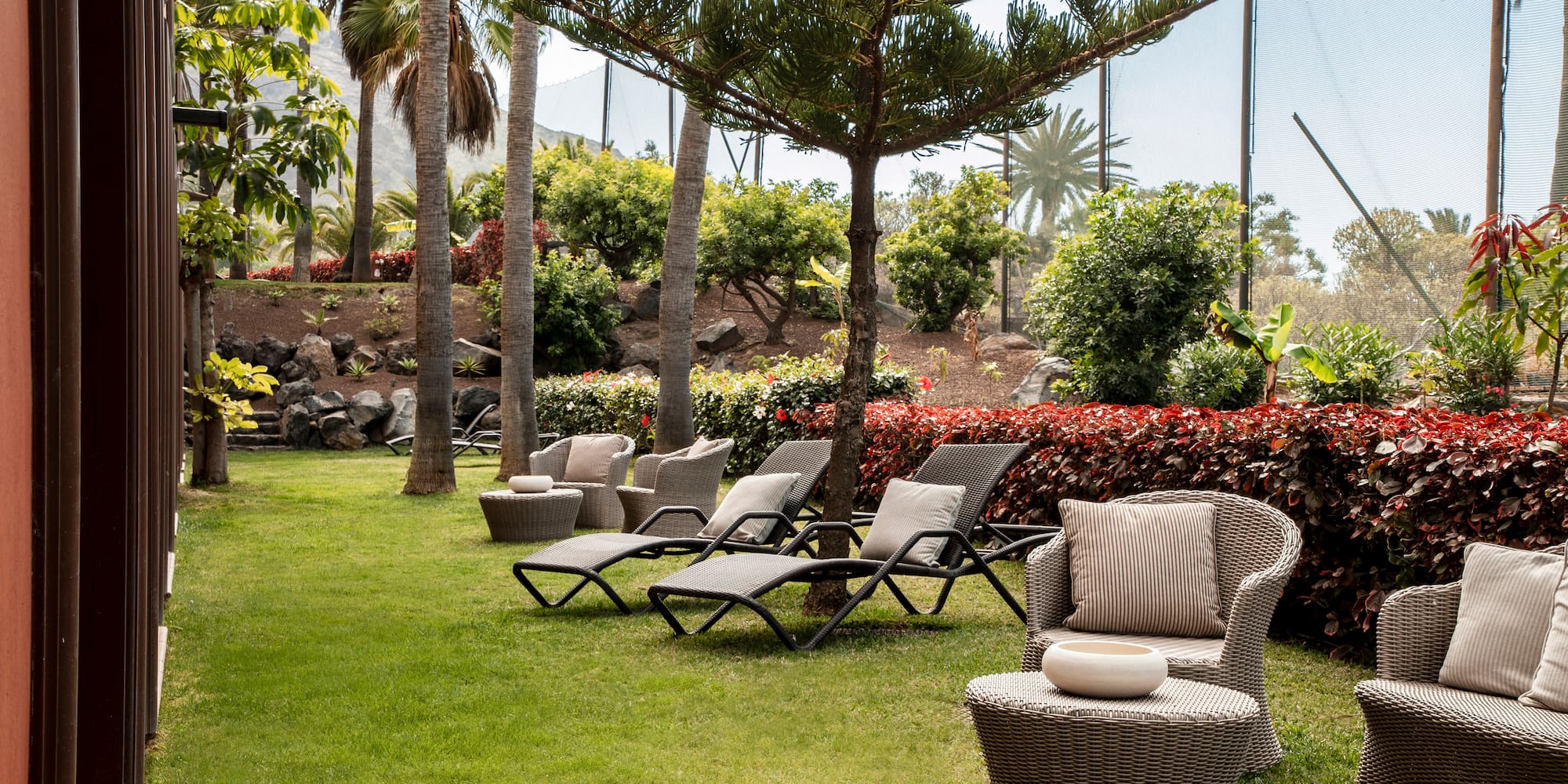 Resort garden lounge area with wicker chairs, palm trees, and lush greenery.