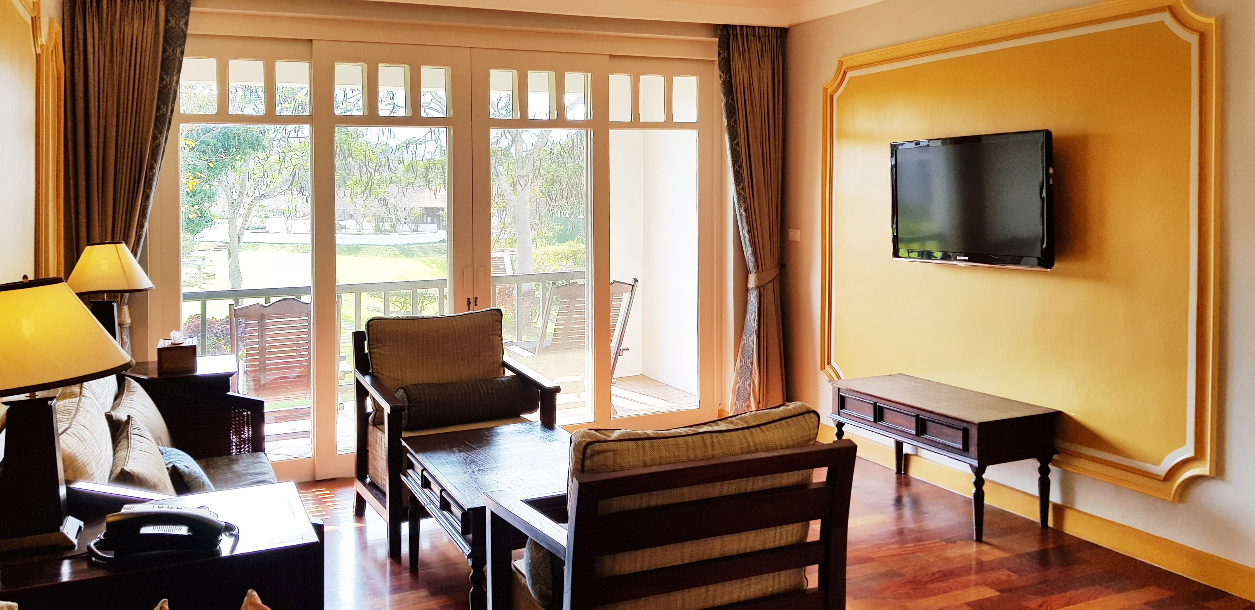 Resort suite with balcony and garden view, wooden chairs, TV on accent wall.