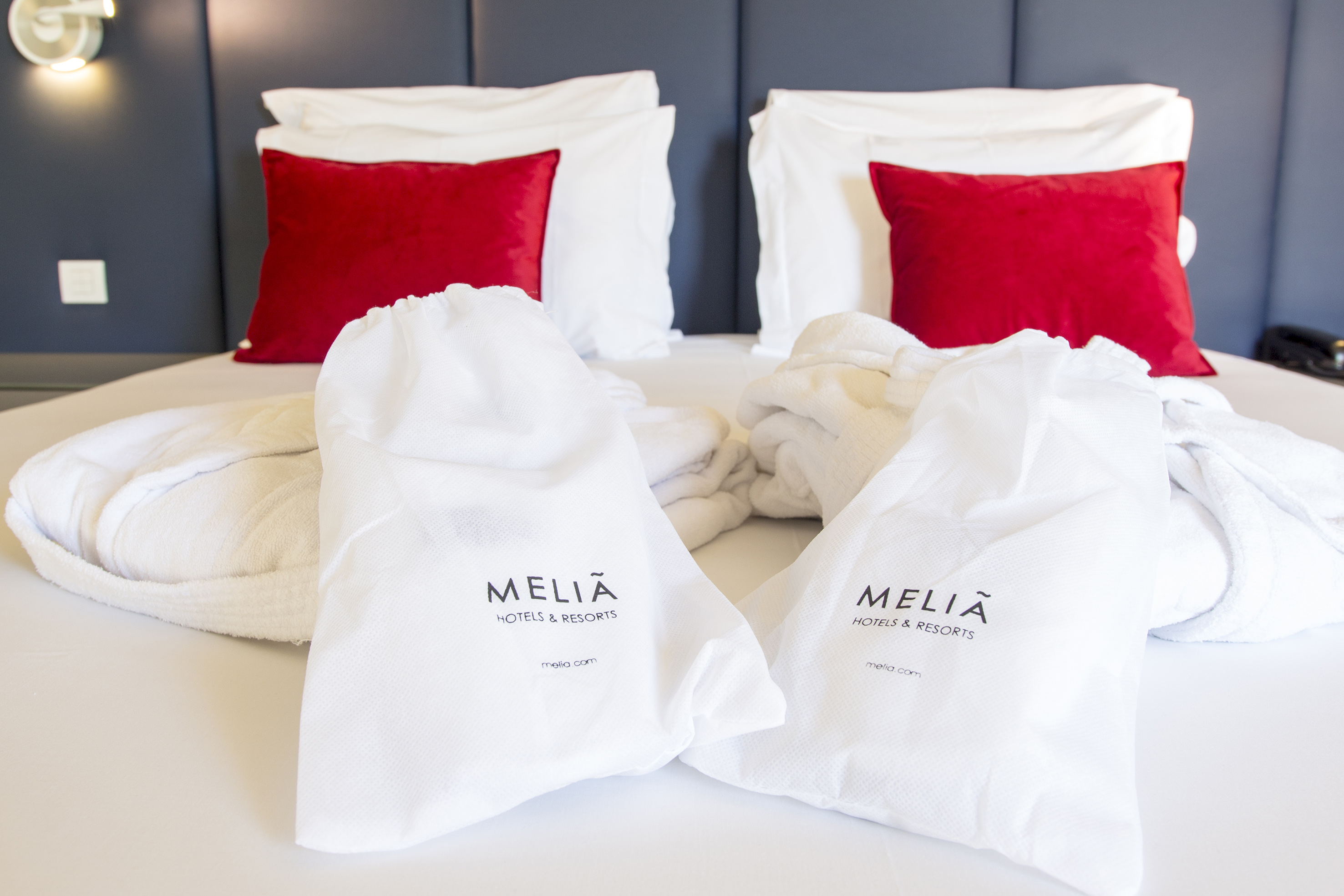 Meliã hotel bed with white bathrobes, red pillows, and branded amenity bags. - Deluxe Room - Meliá Setúbal, Setúbal