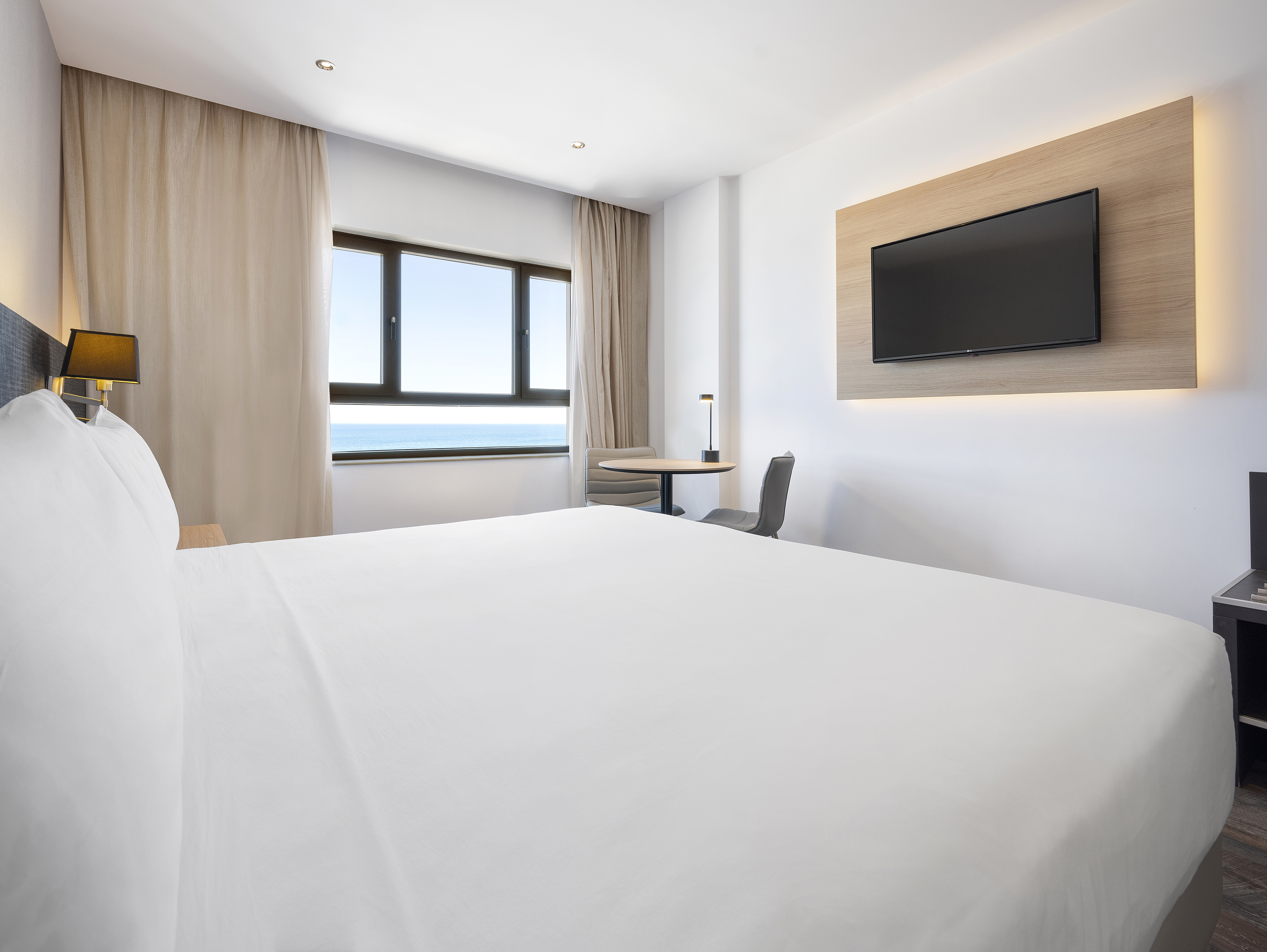 Modern hotel room with white bed, ocean view window, and wall-mounted TV.