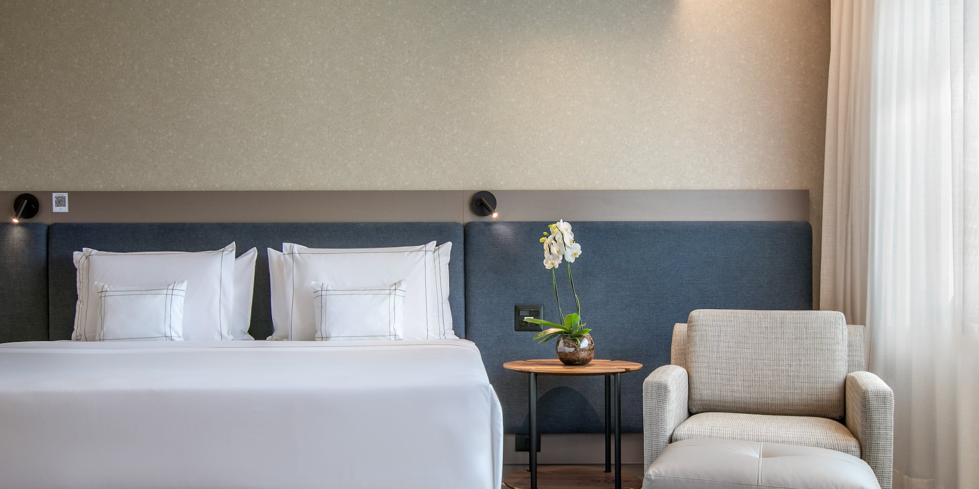 Modern hotel room with bed, armchair, side table, and white orchid.