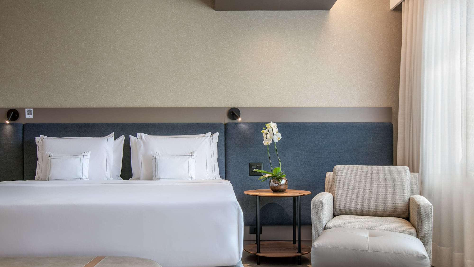 Modern hotel room with bed, armchair, side table, and white orchid.