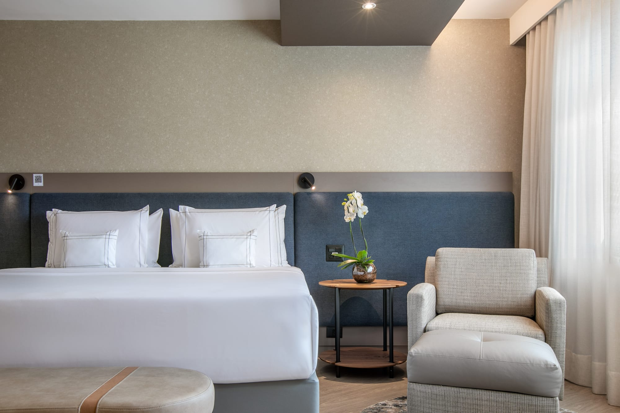 Modern hotel bedroom with large bed, armchair, orchid, and neutral decor.