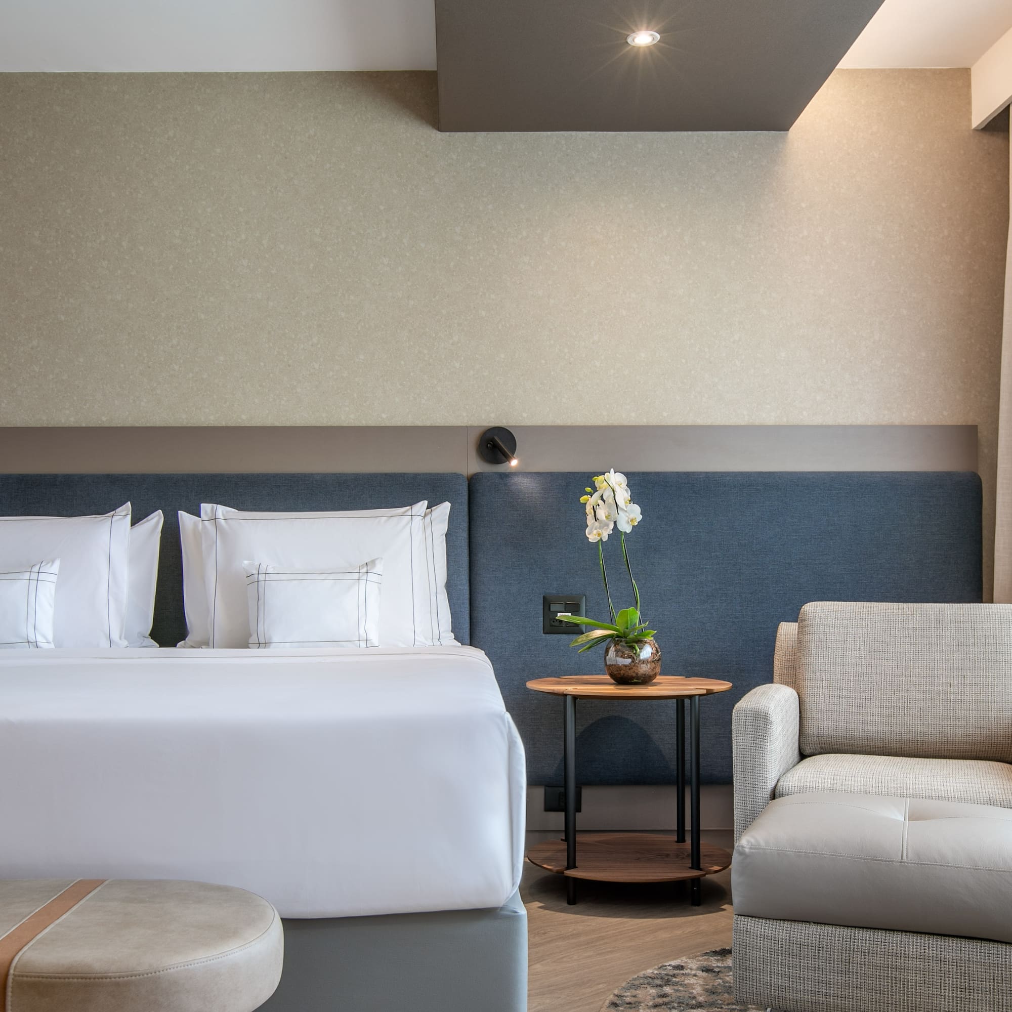 Modern hotel bedroom with large bed, armchair, orchid, and neutral decor.