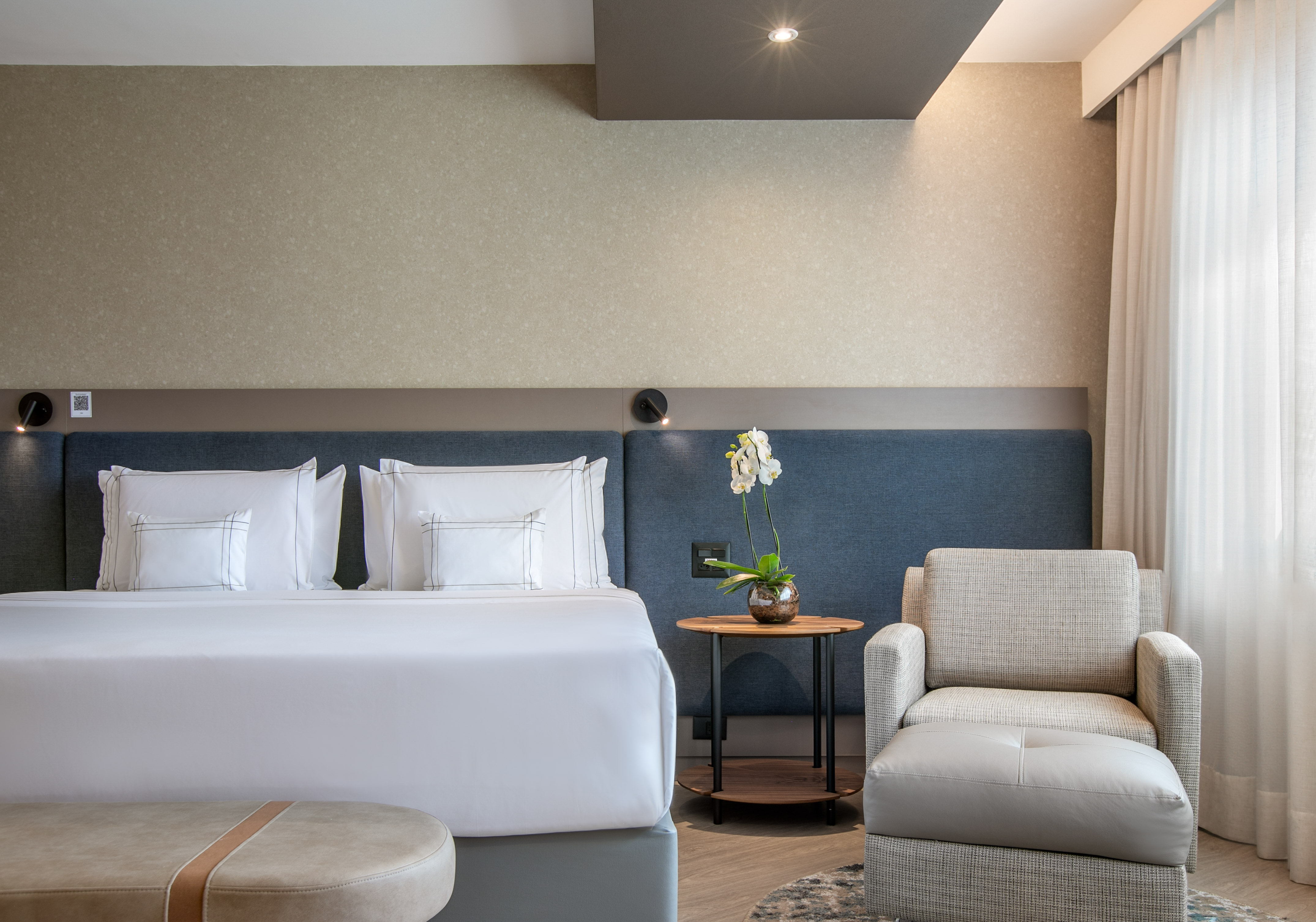 Modern hotel bedroom with large bed, armchair, orchid, and neutral decor.