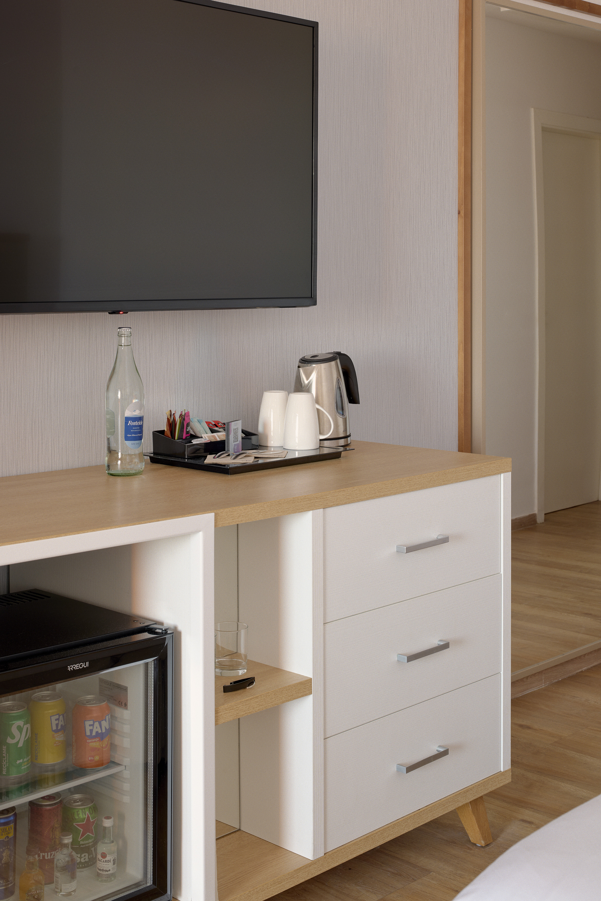 Hotel room with TV, mini-bar fridge, kettle, water, and coffee supplies on dresser.