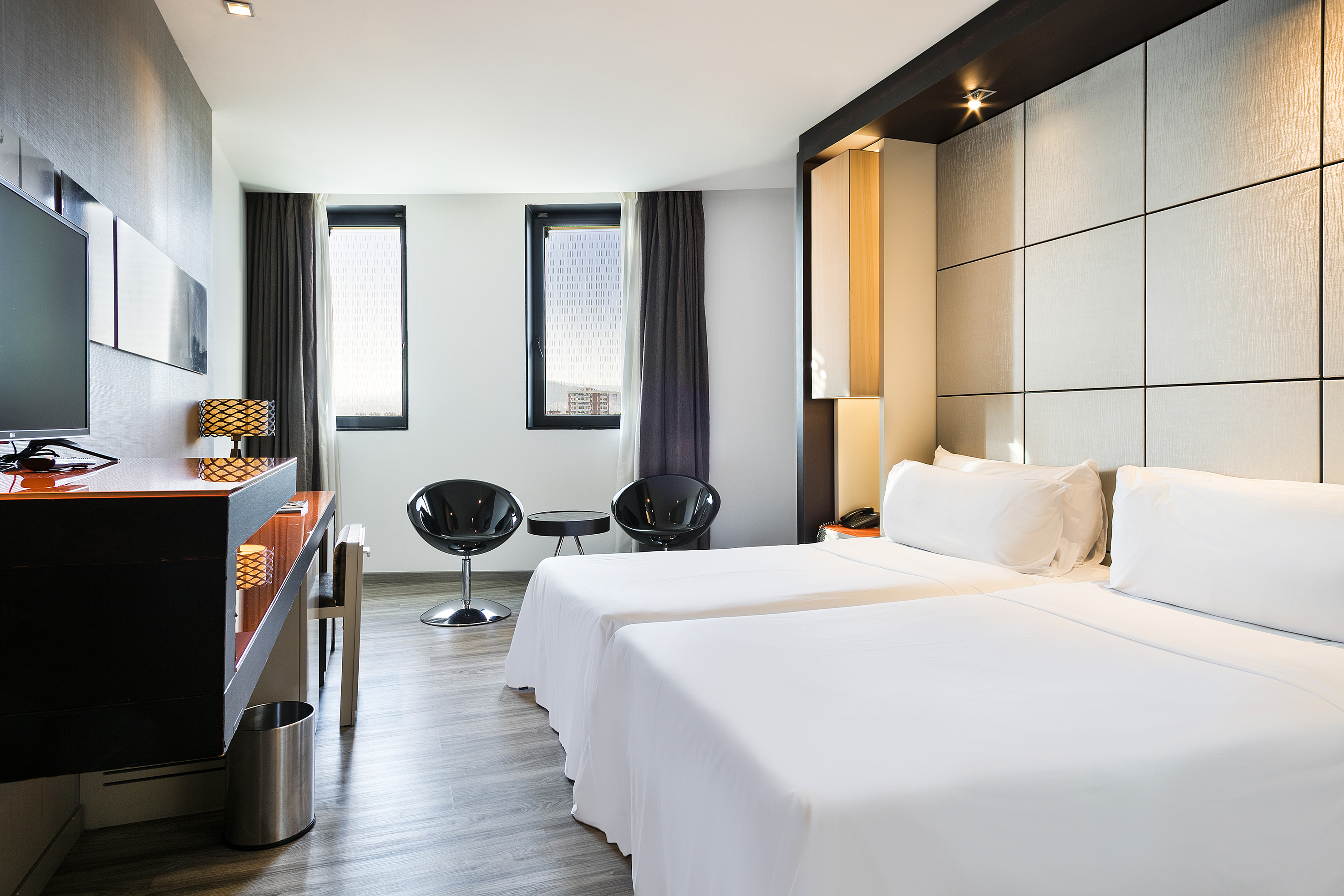 Modern hotel room with two twin beds, desk, TV, and black accent chairs.