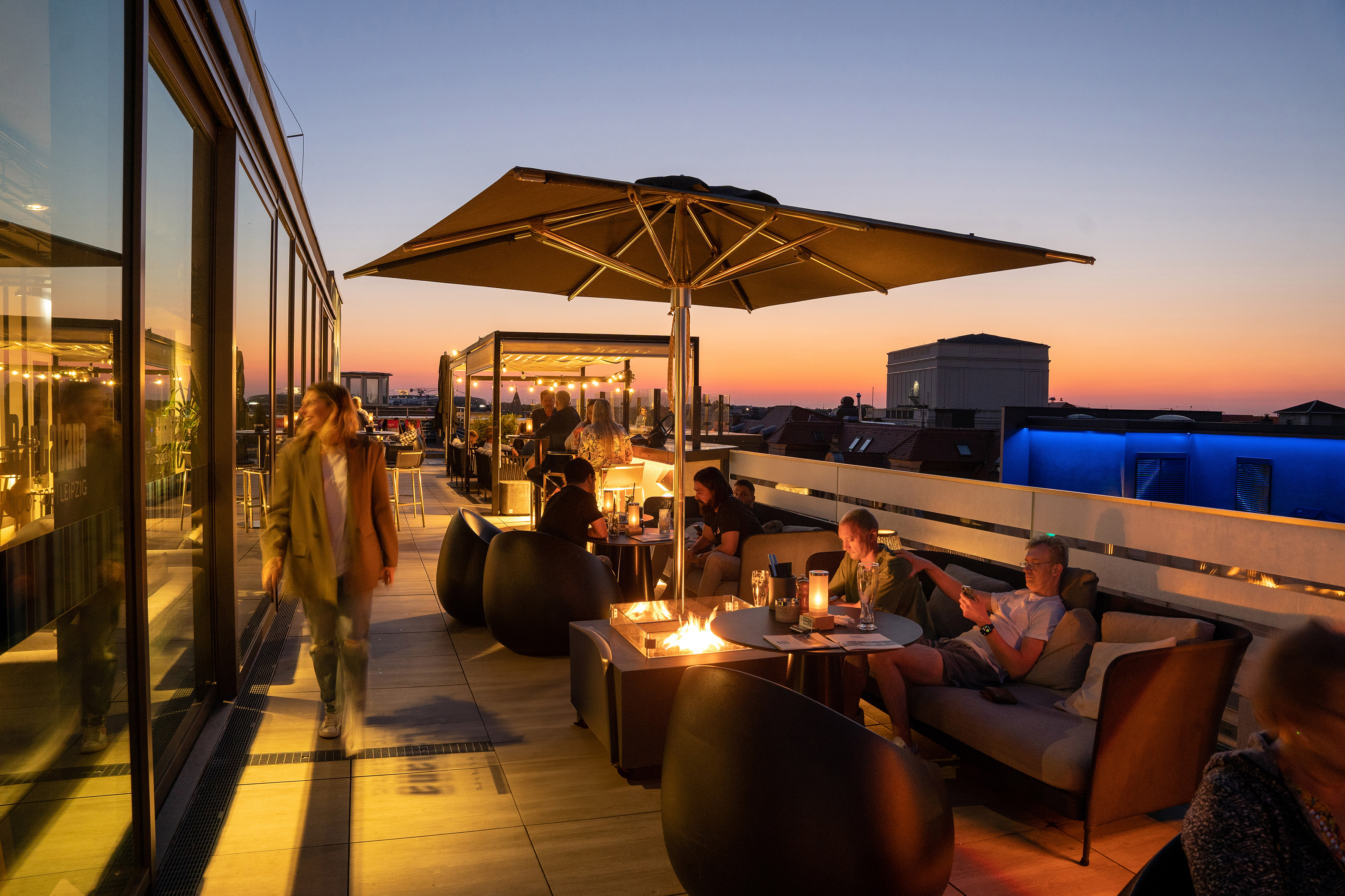 Rooftop bar with people enjoying drinks, a fire pit, and city views at sunset.
