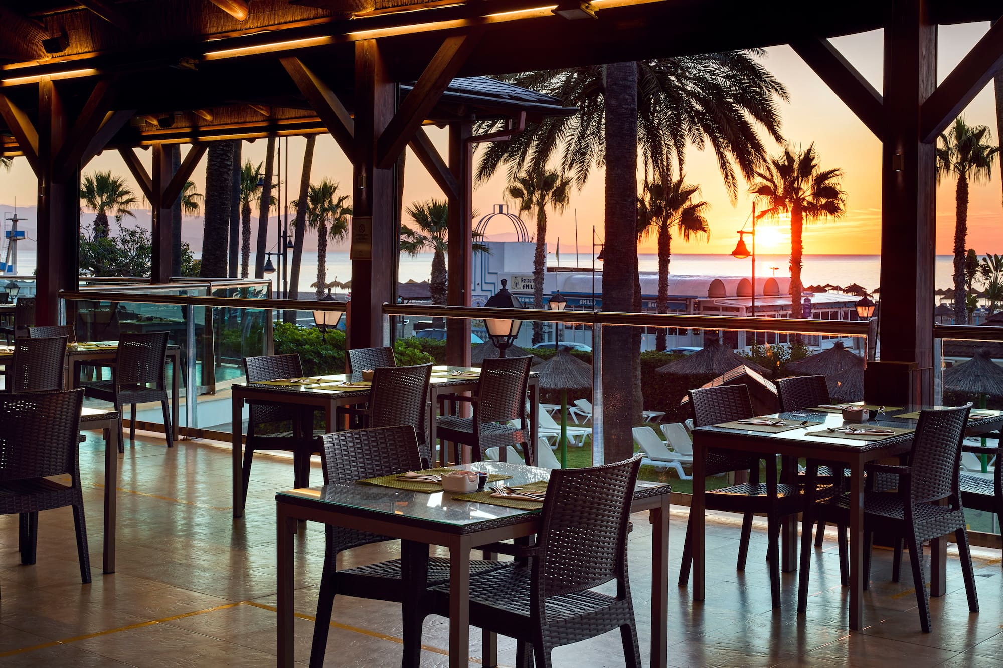 Tropical resort dining terrace with ocean sunset and palm trees.