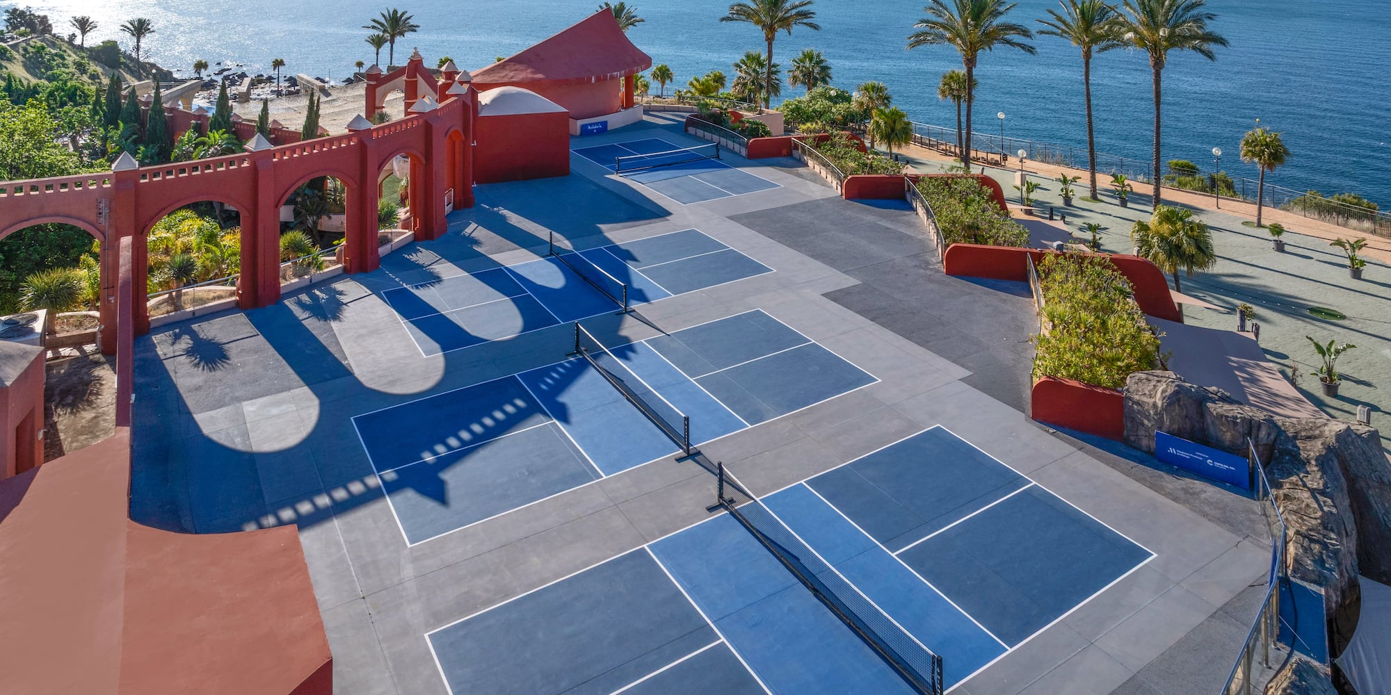 Blue pickleball courts by the ocean, with red archways and palm trees.