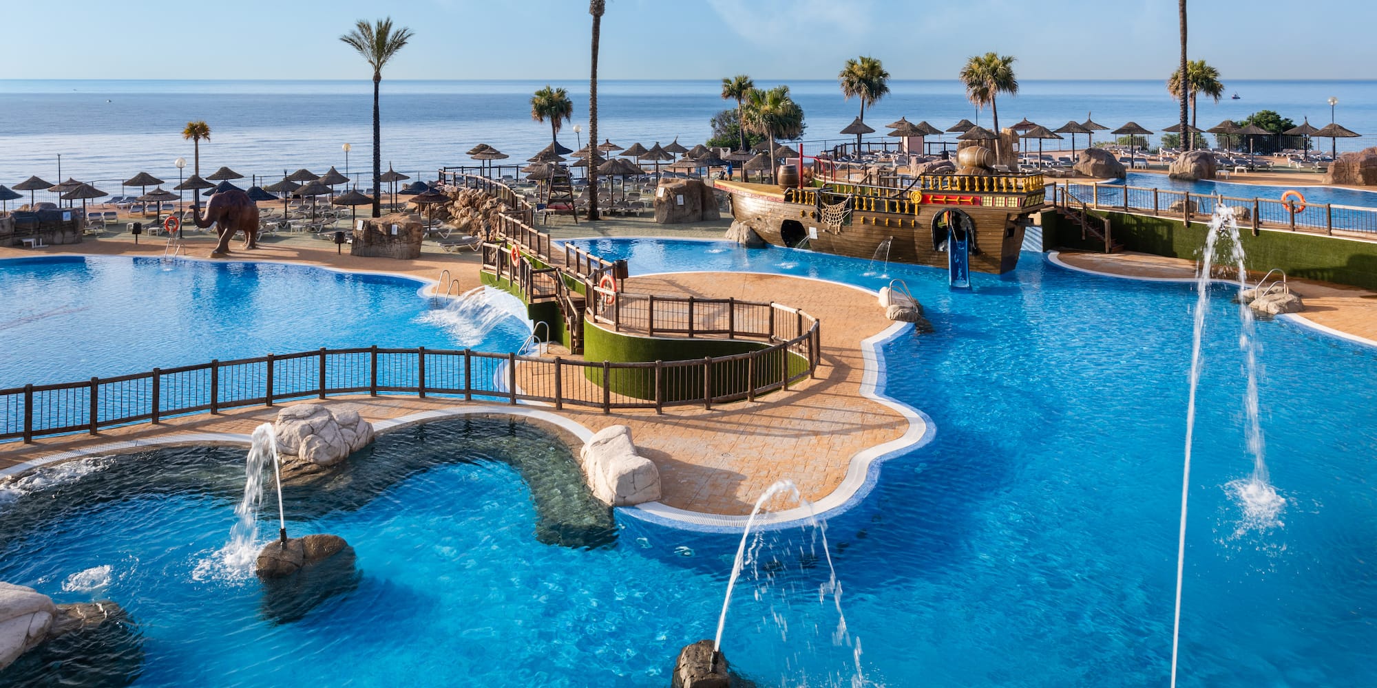 Resort swimming pool with pirate ship, palm trees, beach, and ocean view.