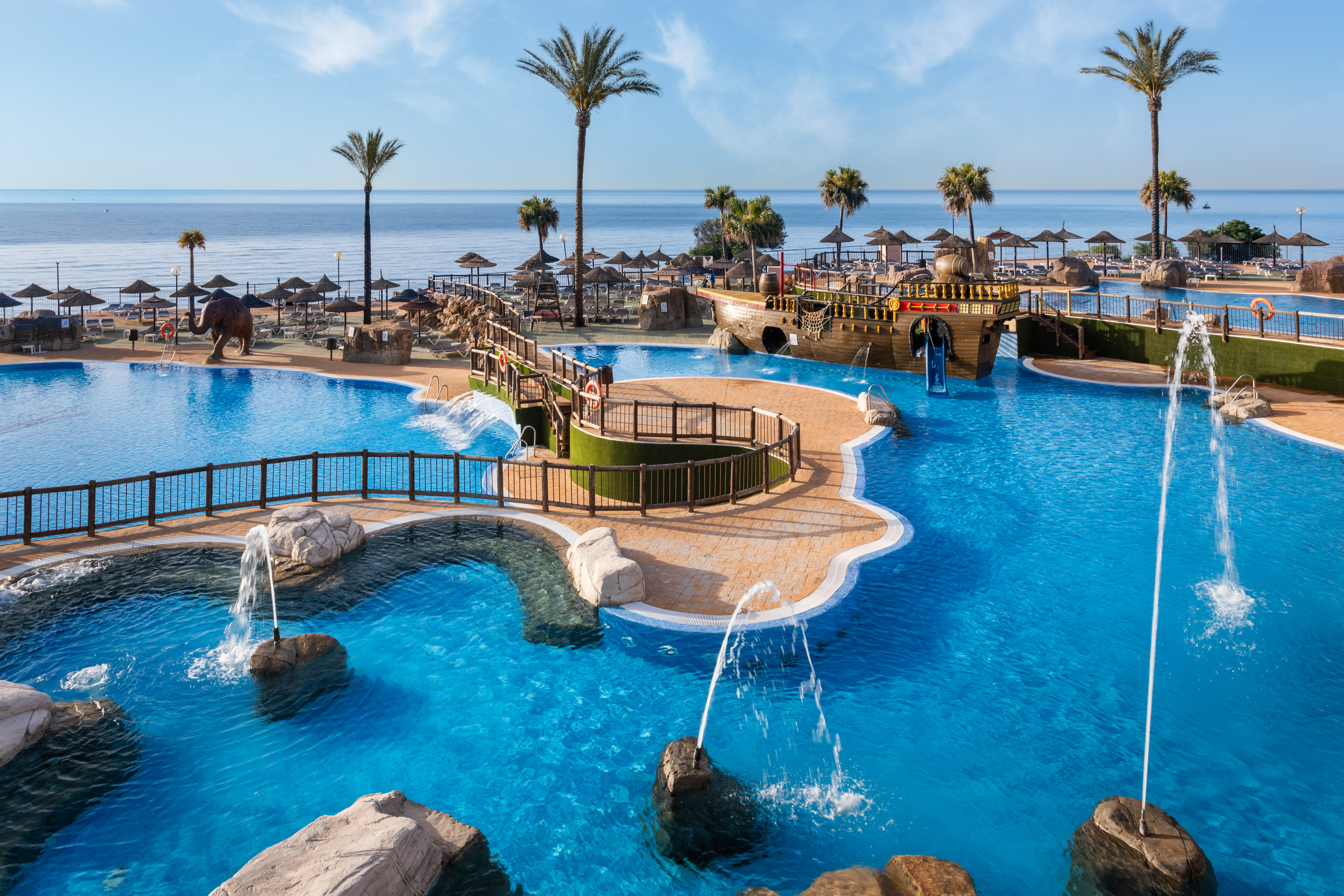 Resort swimming pool with pirate ship, palm trees, beach, and ocean view.