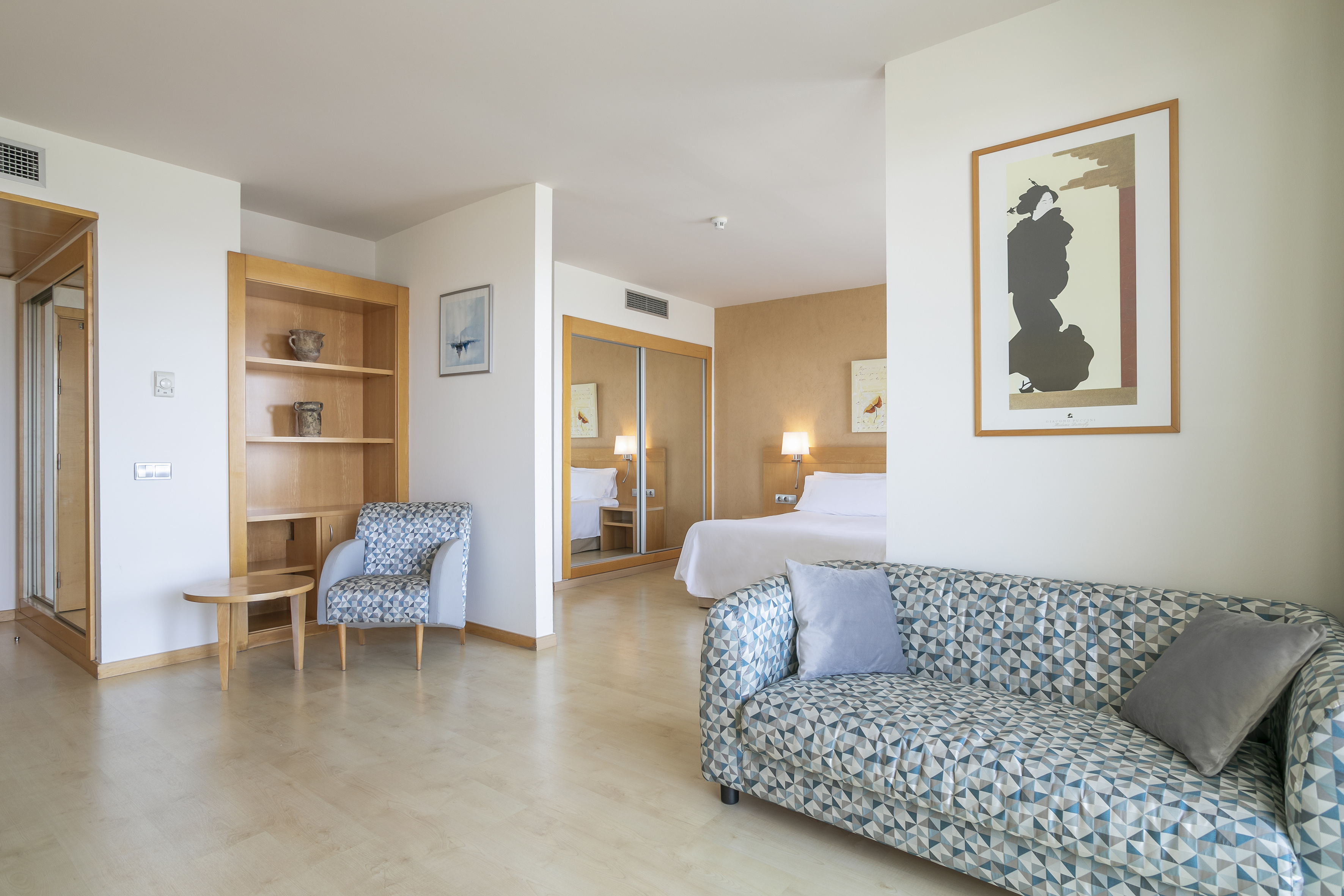 Hotel suite with patterned sofa, wooden bookshelf, and bedroom beyond.