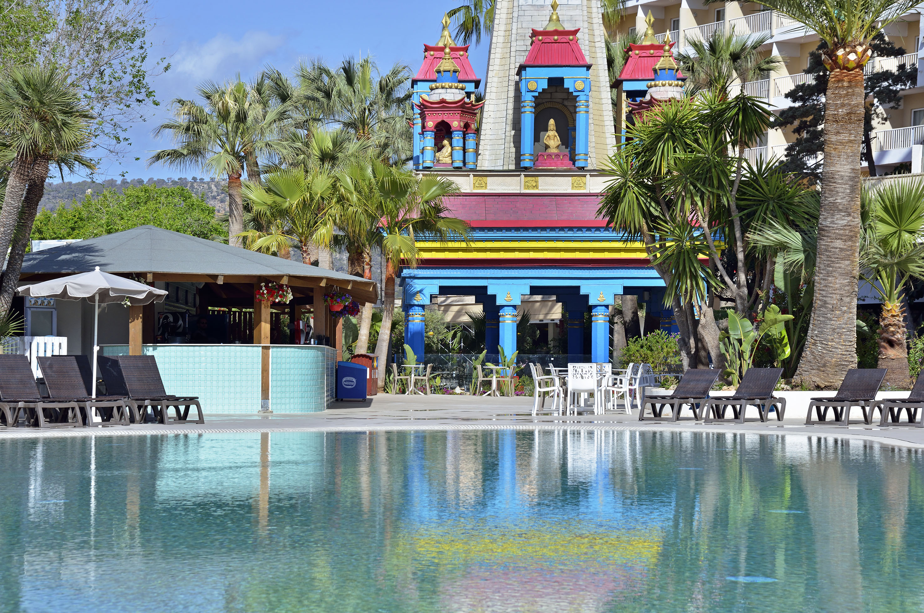 Tropical resort pool with colorful building, palm trees, and sun loungers.