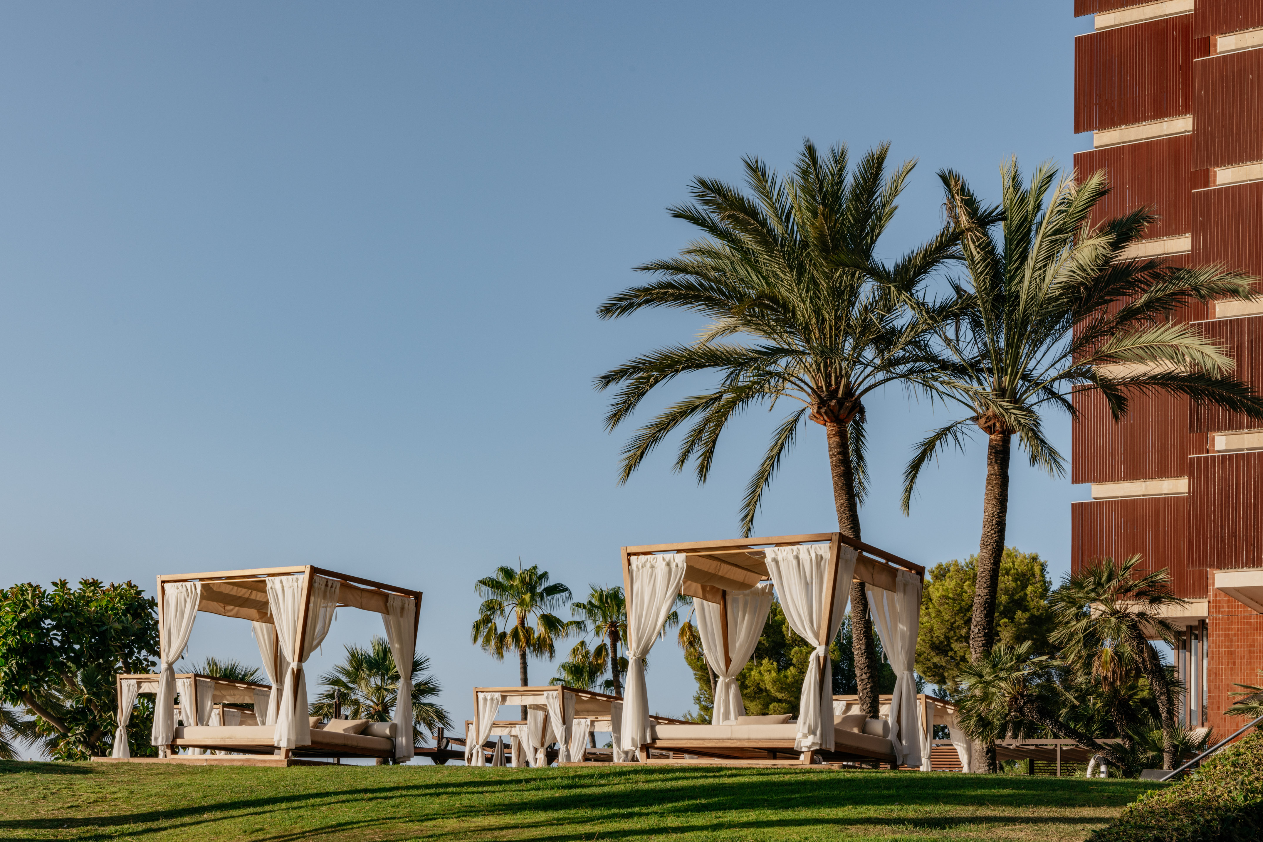 Sunny resort garden with wooden cabanas, palm trees, and hotel facade.