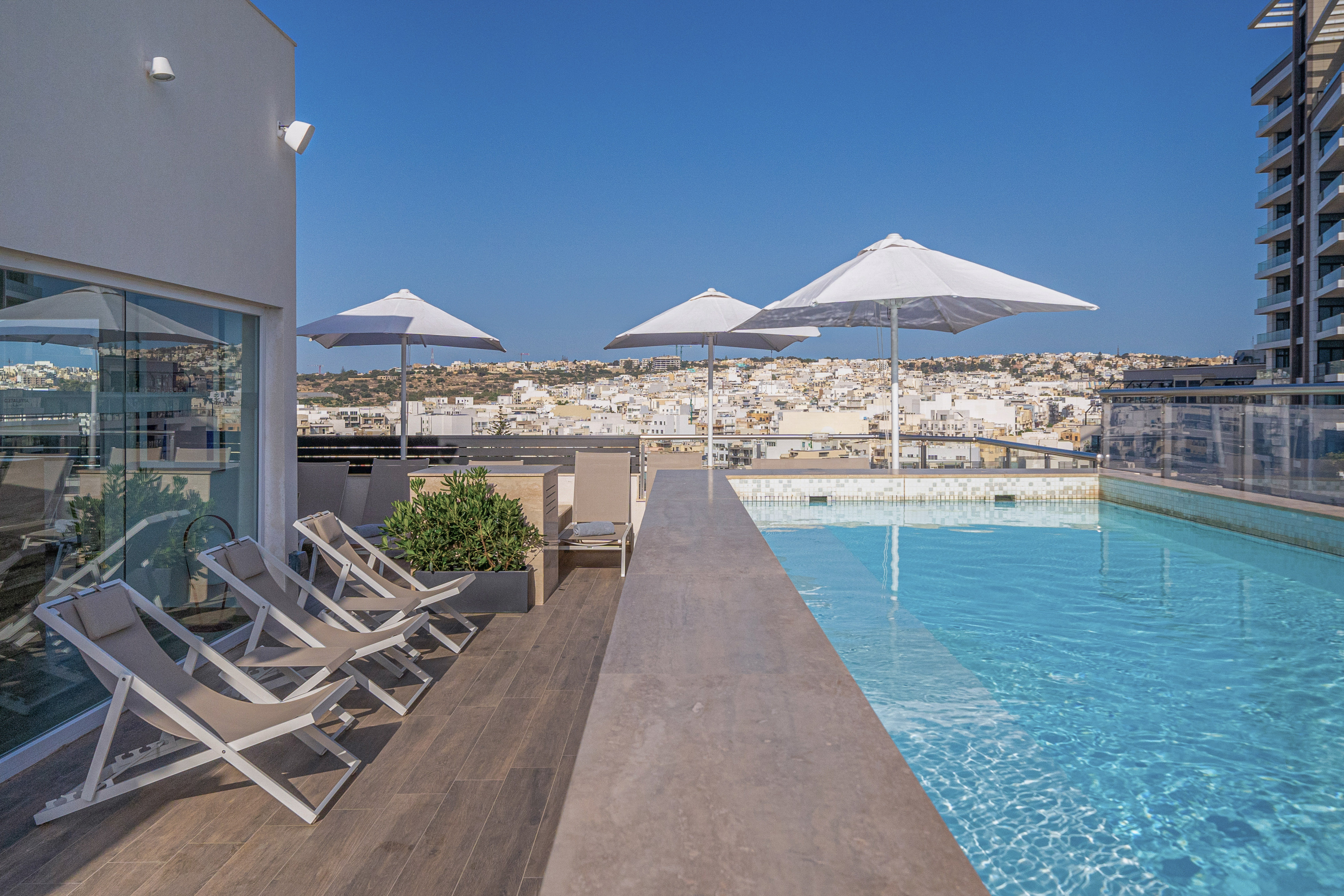 Rooftop infinity pool deck with lounge chairs, white umbrellas, and city views.
