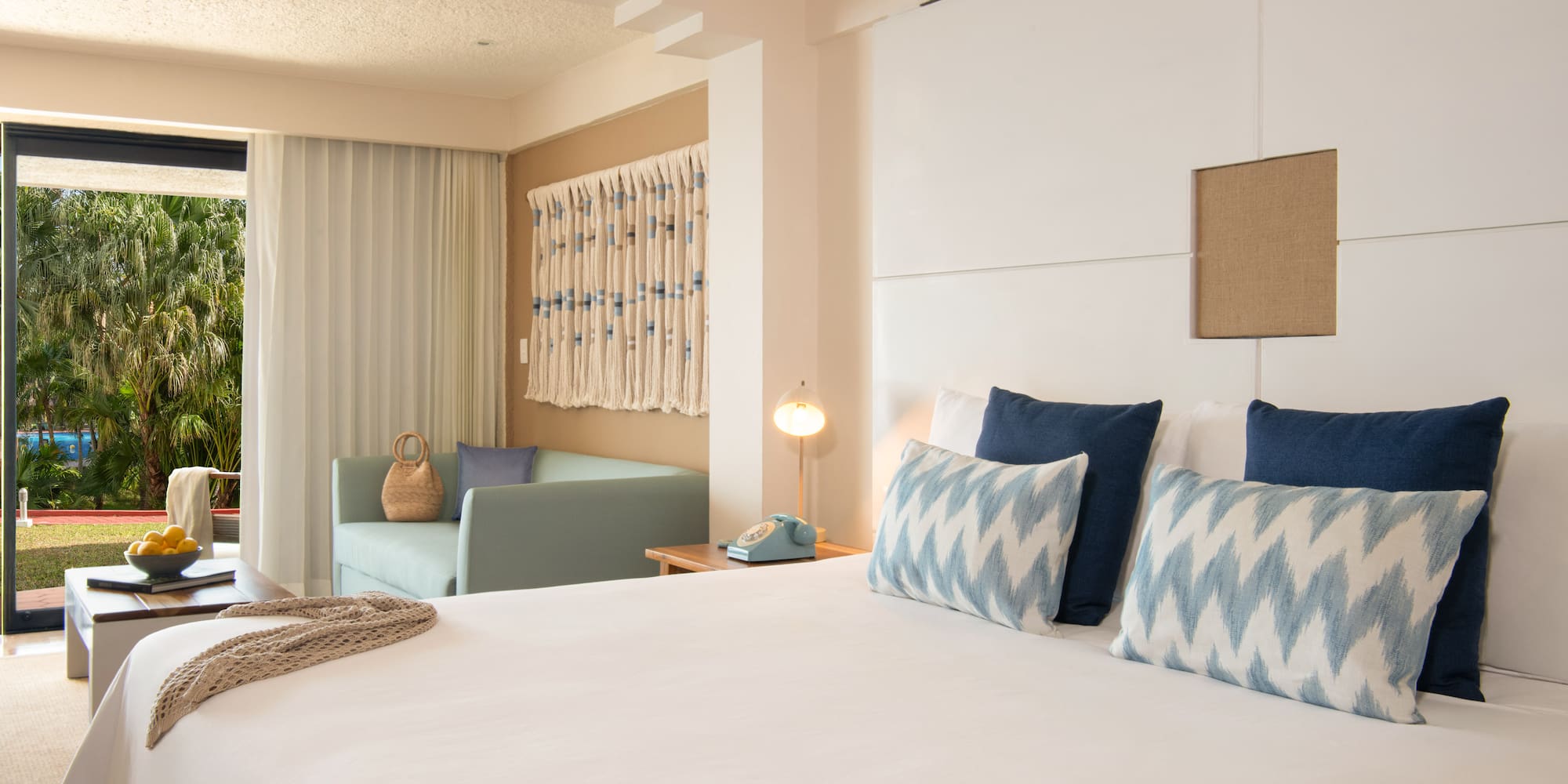 Hotel room with bed, blue pillows, sofa, and tropical palm garden view.