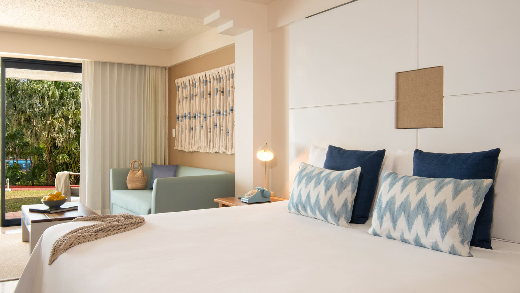 Hotel room with bed, blue pillows, sofa, and tropical palm garden view.